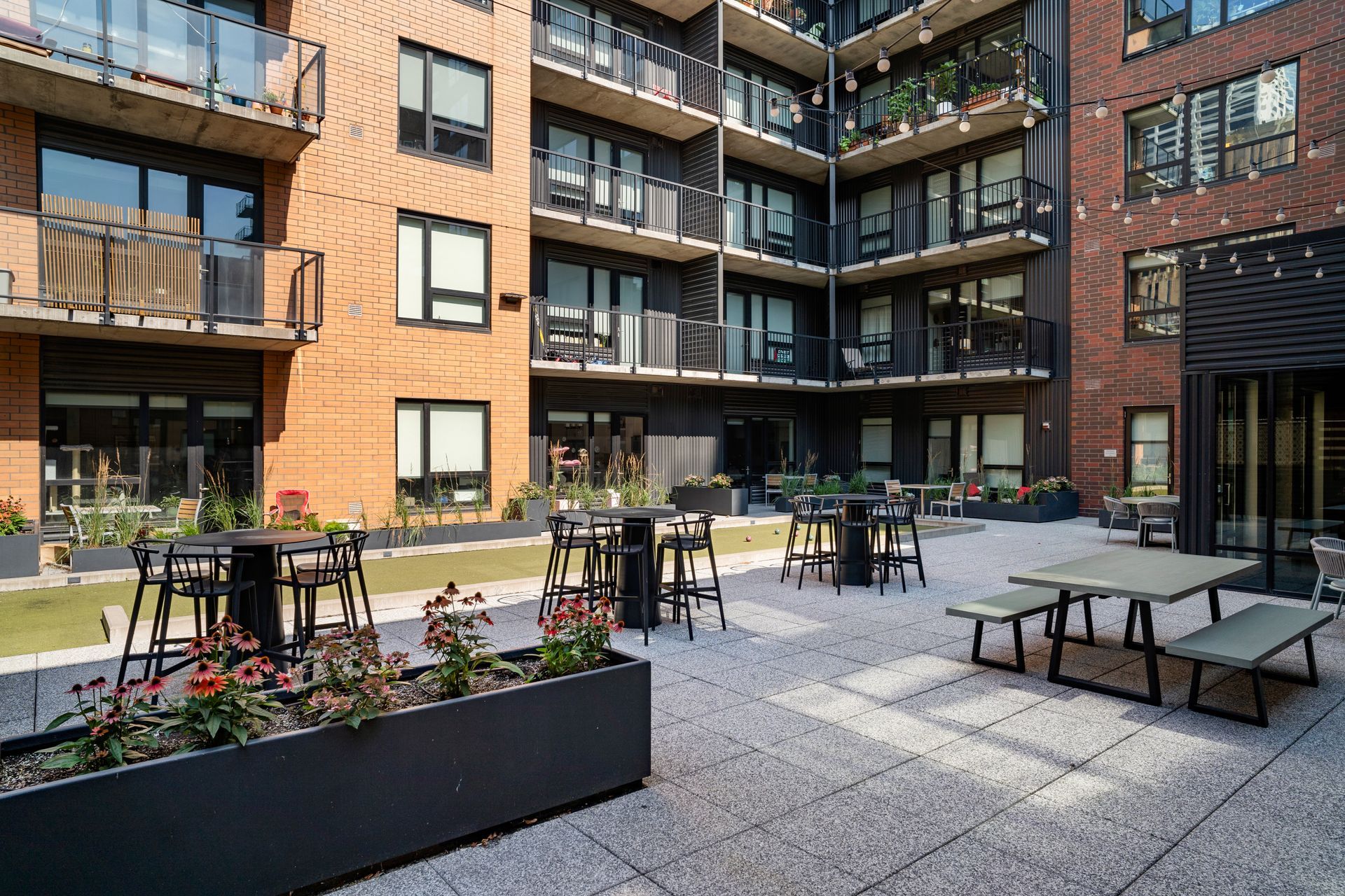 Outdoor patio with seating at an apartment complex. Brick building, black tables, gray pavement, and flower boxes.