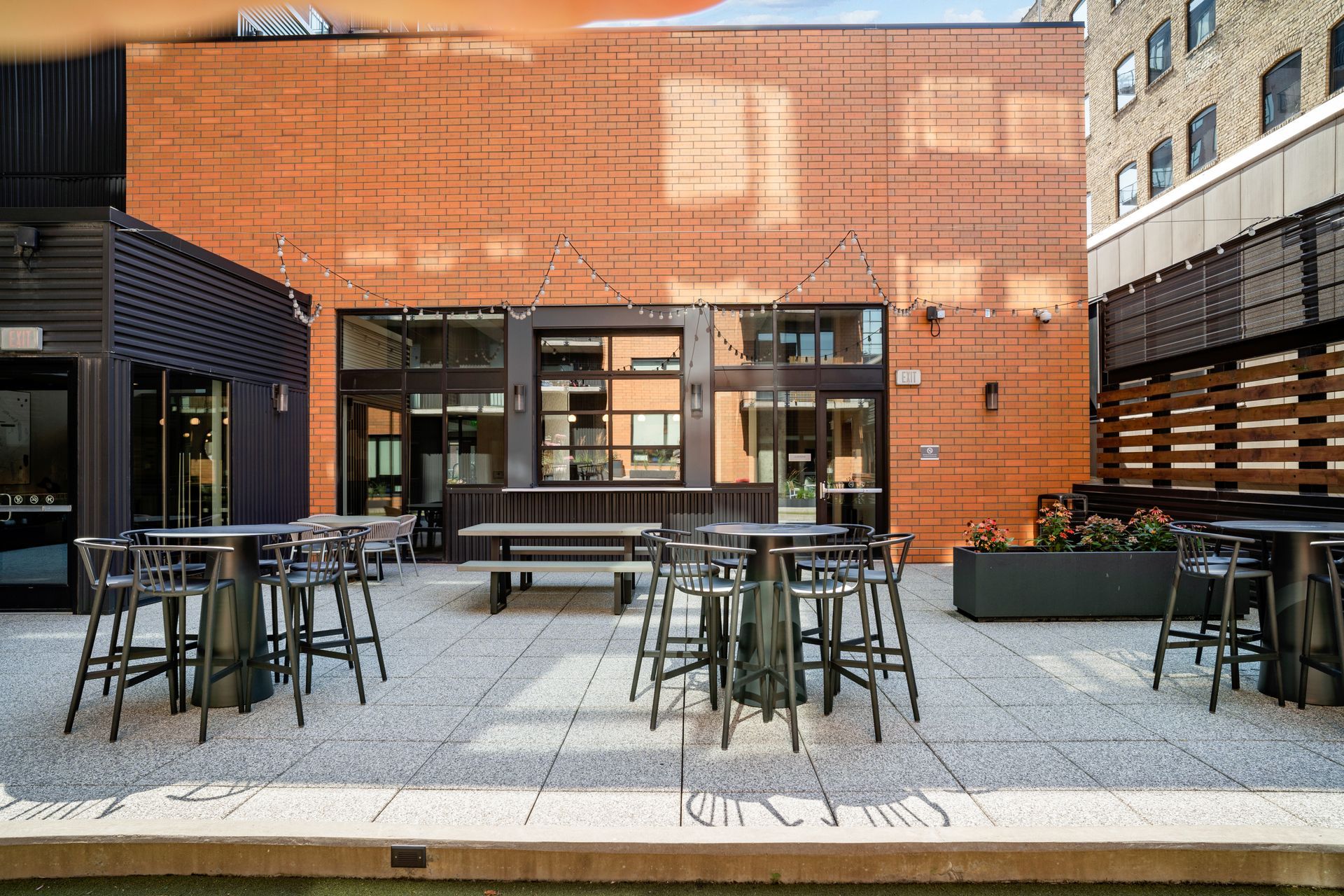 Outdoor patio with bar tables and chairs, brick wall backdrop. String lights above.