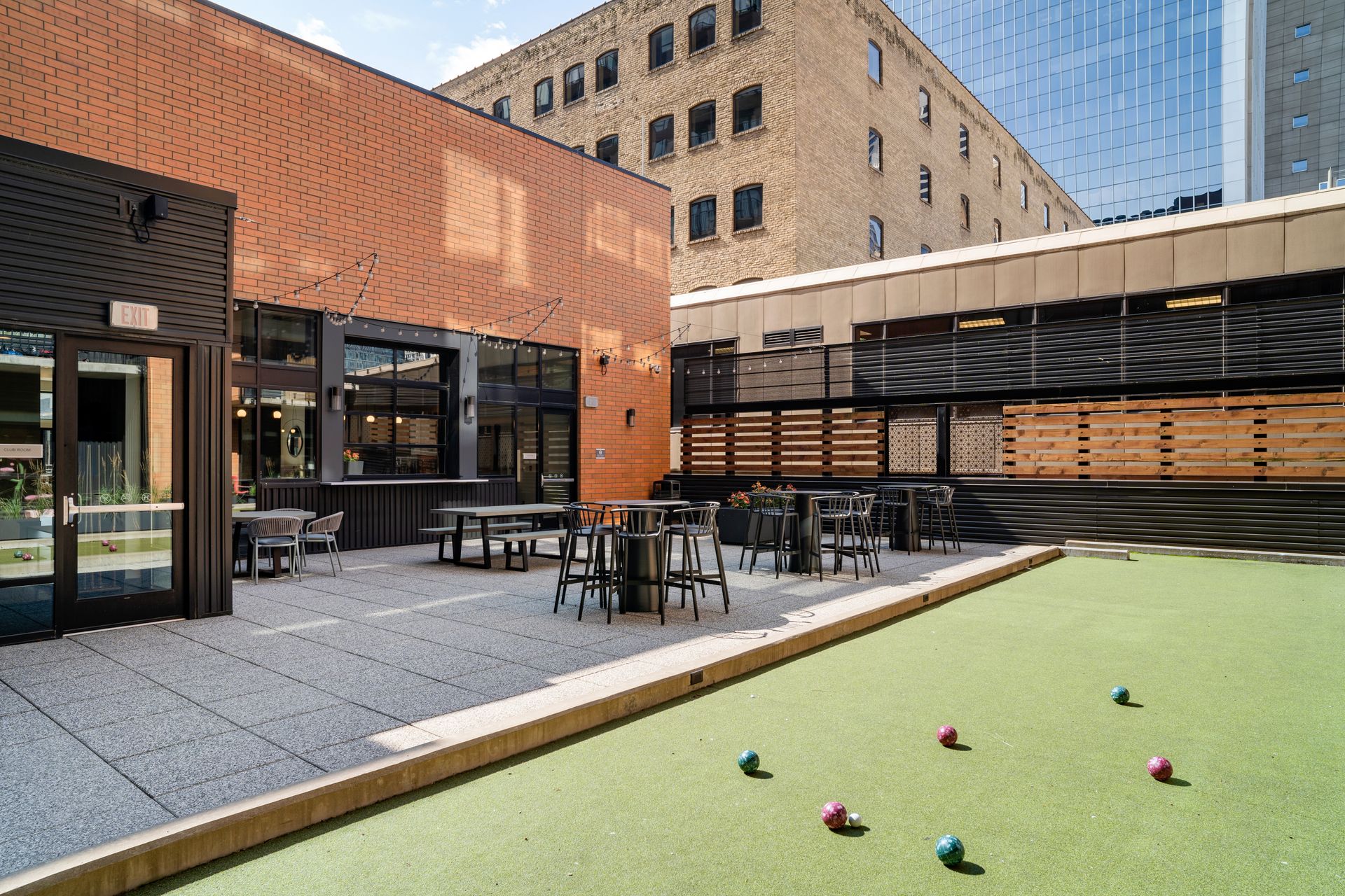 Outdoor patio with bocce balls on artificial turf. Tables, chairs, and brick building in background.