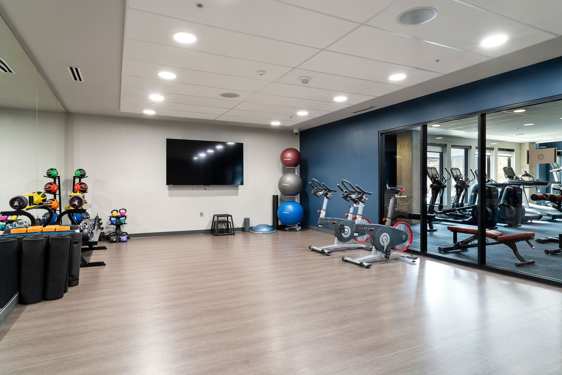 A fitness center with exercise equipment, medicine balls, a TV, and a view into another gym space.