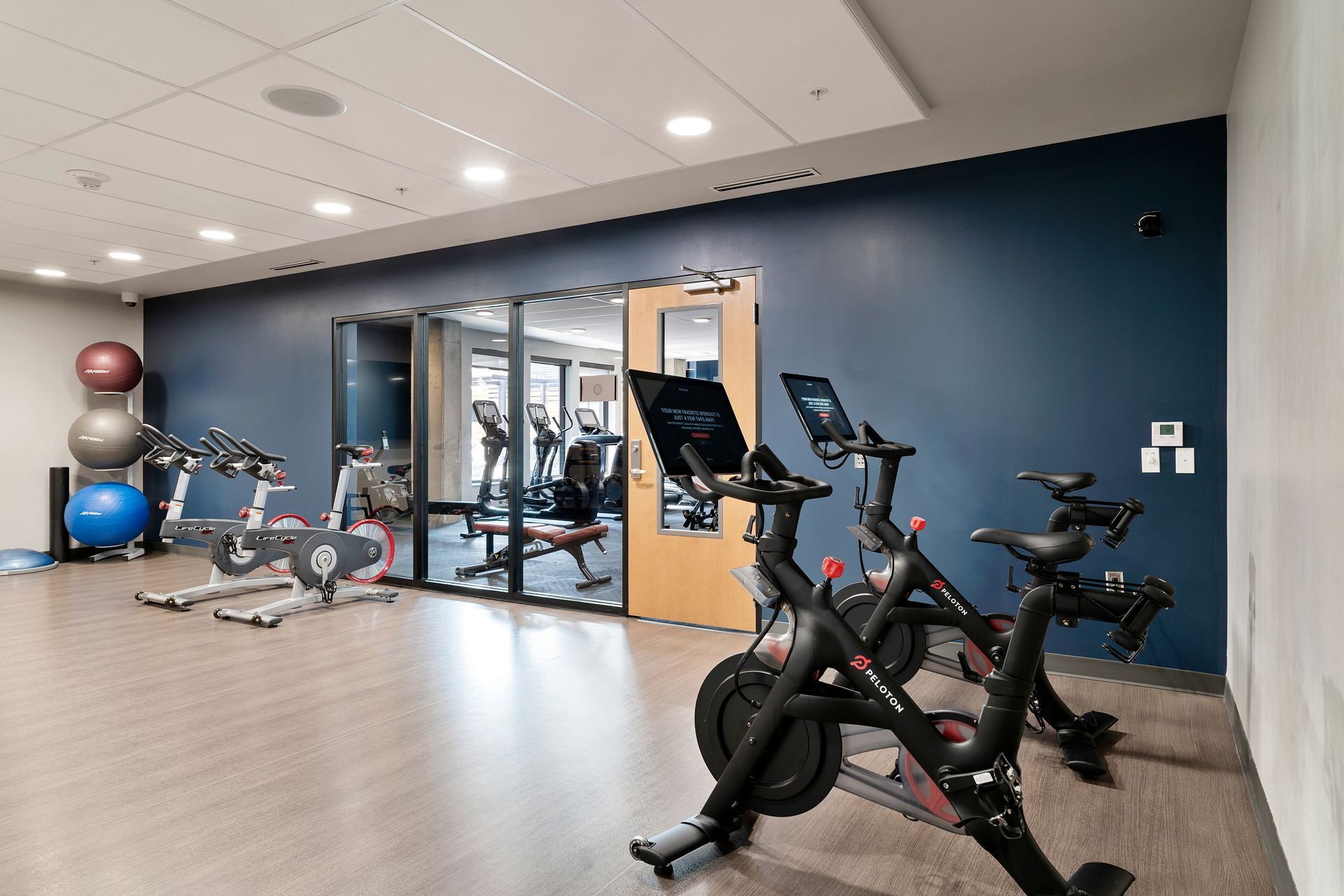 Gym with spin bikes, exercise balls, and a view of a second gym area through glass doors. Blue wall.