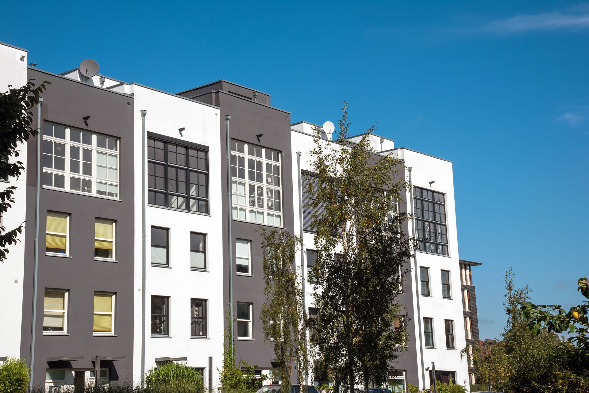 Modern multi-story apartment building with white and gray facades, trees, and blue sky.