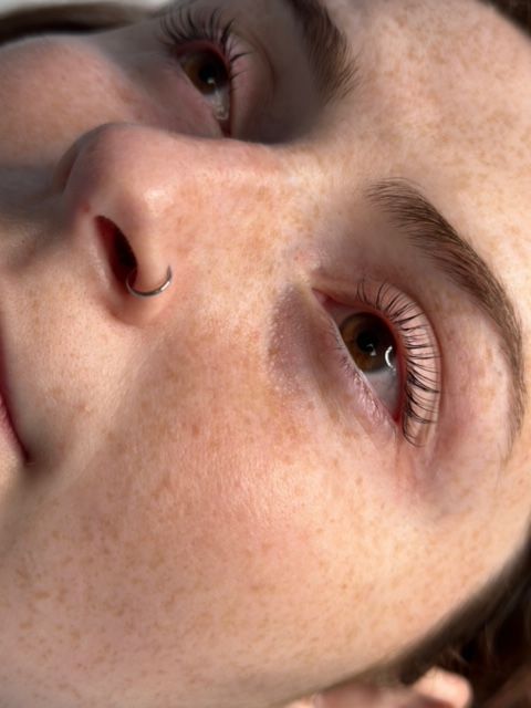 Close-up of a person's face with freckles, a nose ring, and curled eyelashes looking upward.
