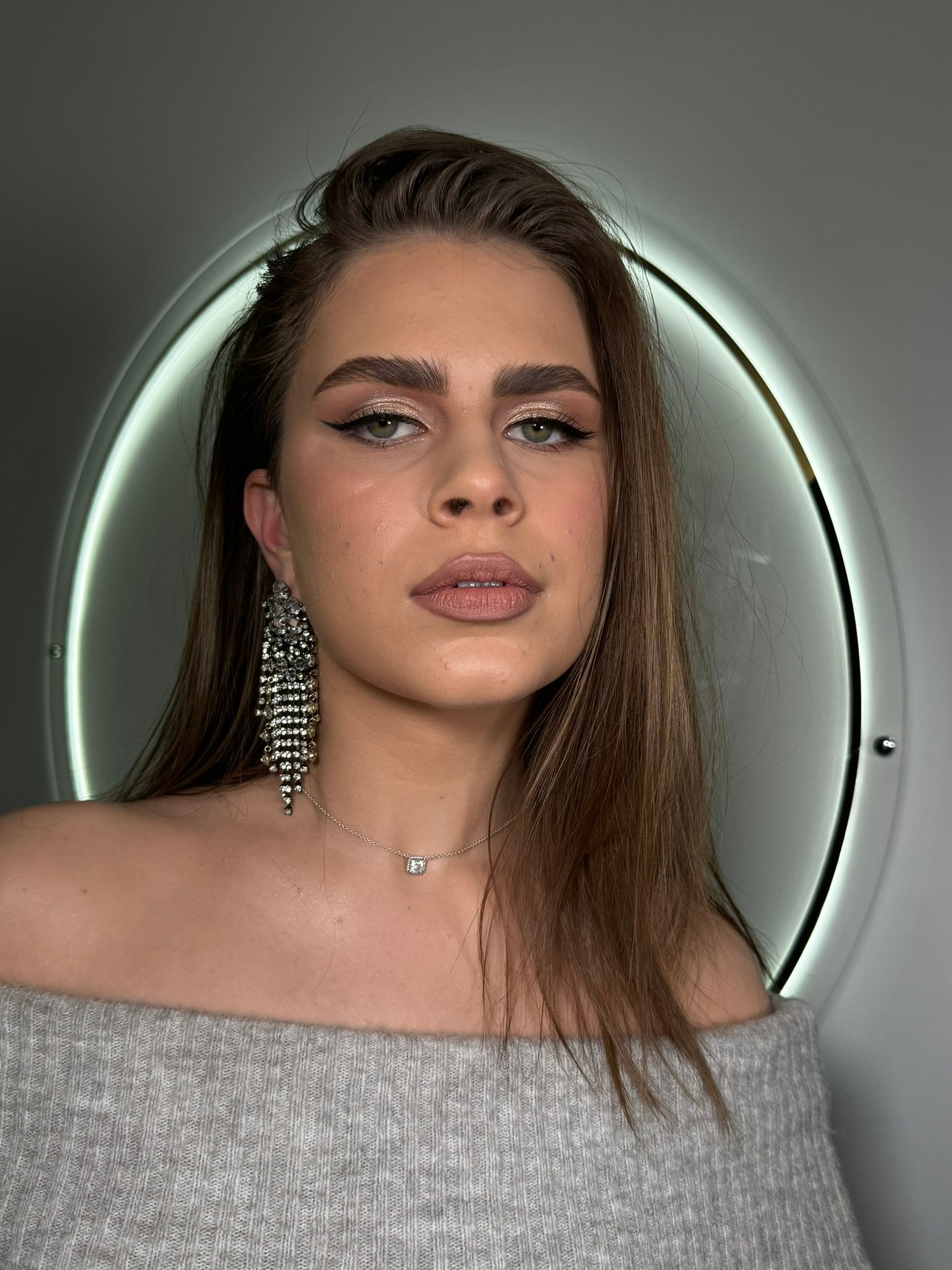 Woman with glamorous makeup and jewelry, gray sweater. She is in front of a circular, illuminated mirror.