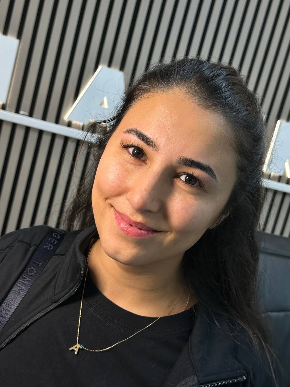 Woman with dark hair smiles, wearing a black jacket and a gold necklace.  Background shows a wall.