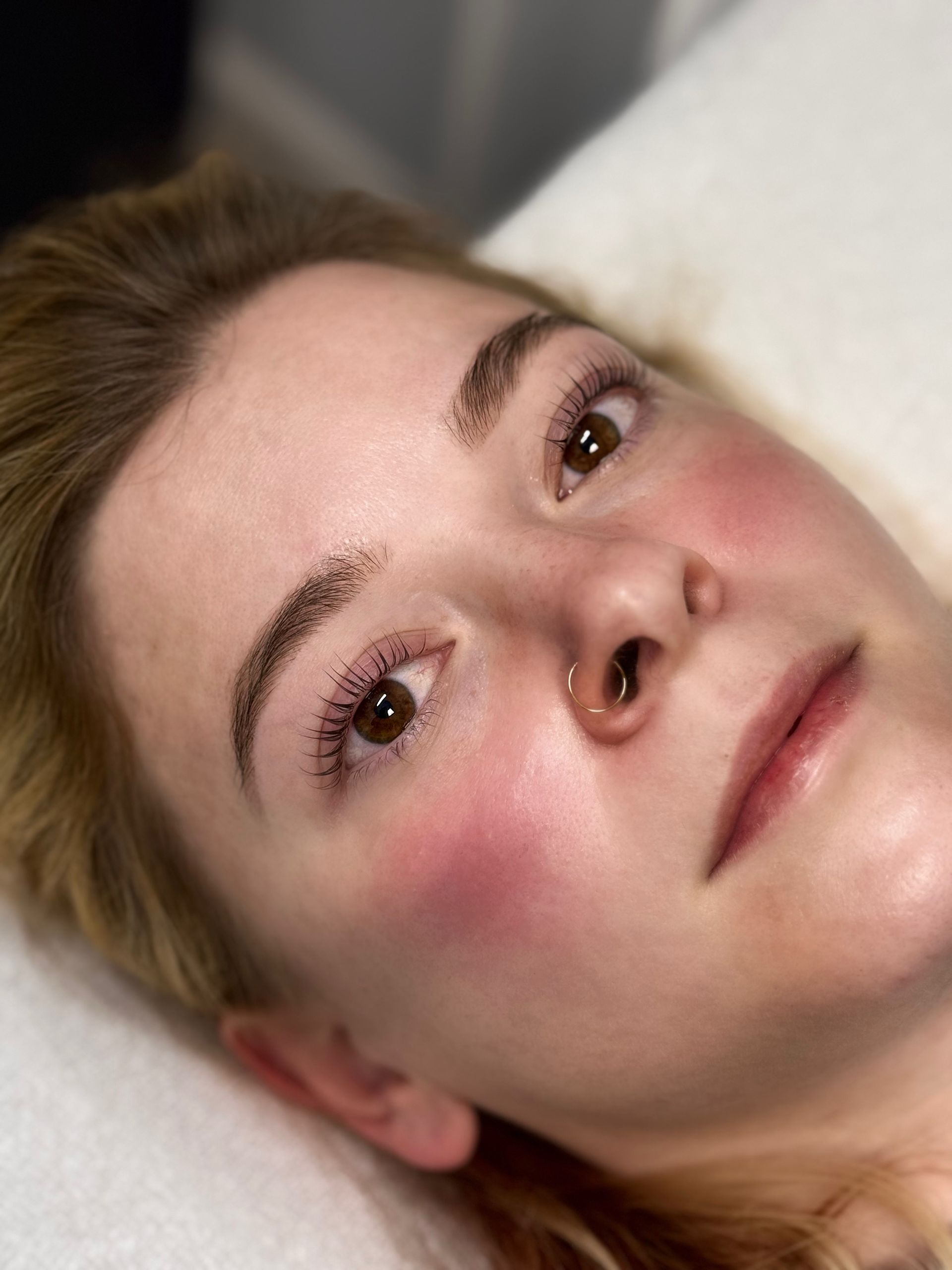 Woman with semi-permanent eyebrow tattoo and reddened cheeks, lying on a white surface.
