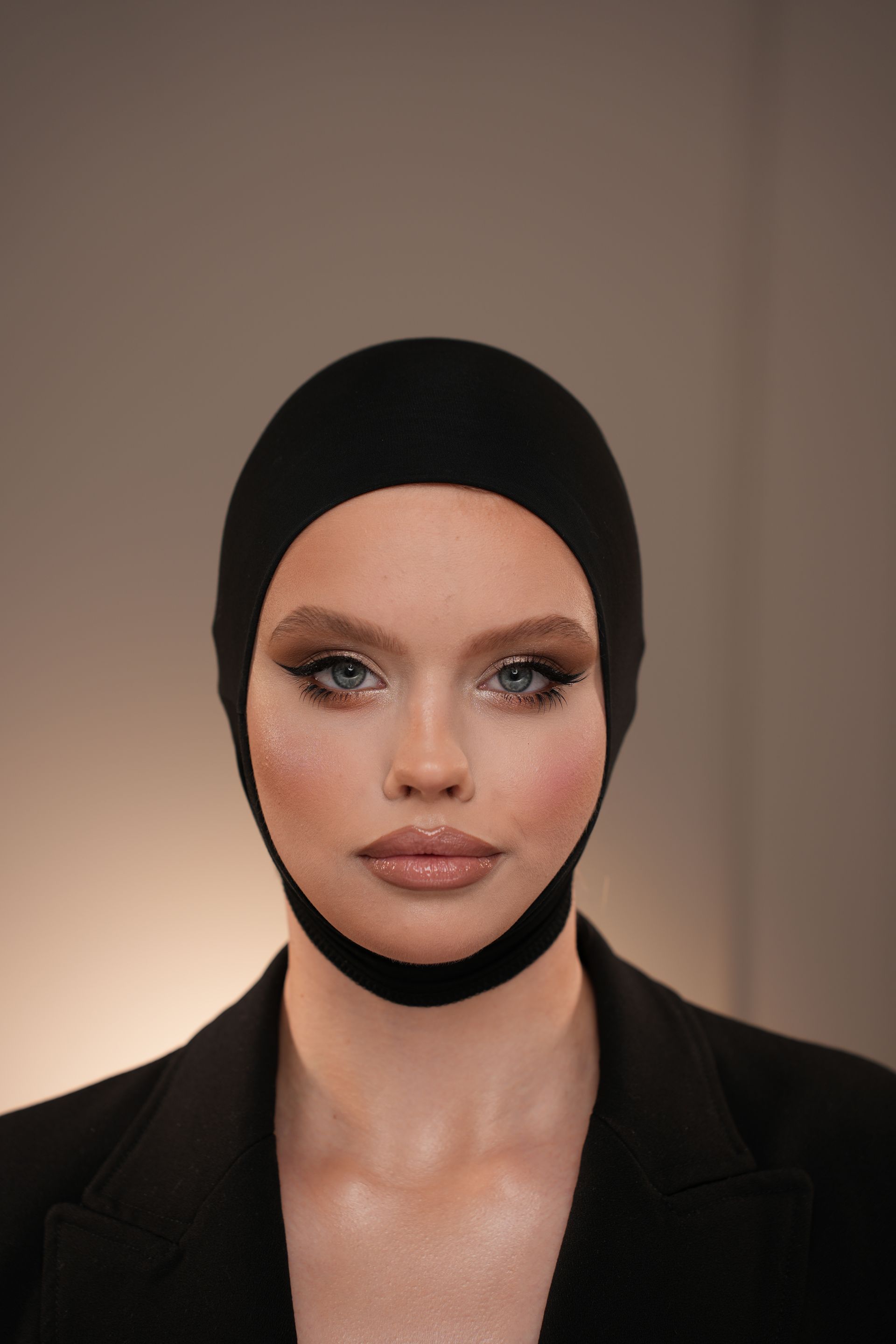 Woman wearing a black head covering and blazer, with dramatic eye makeup and neutral lips.