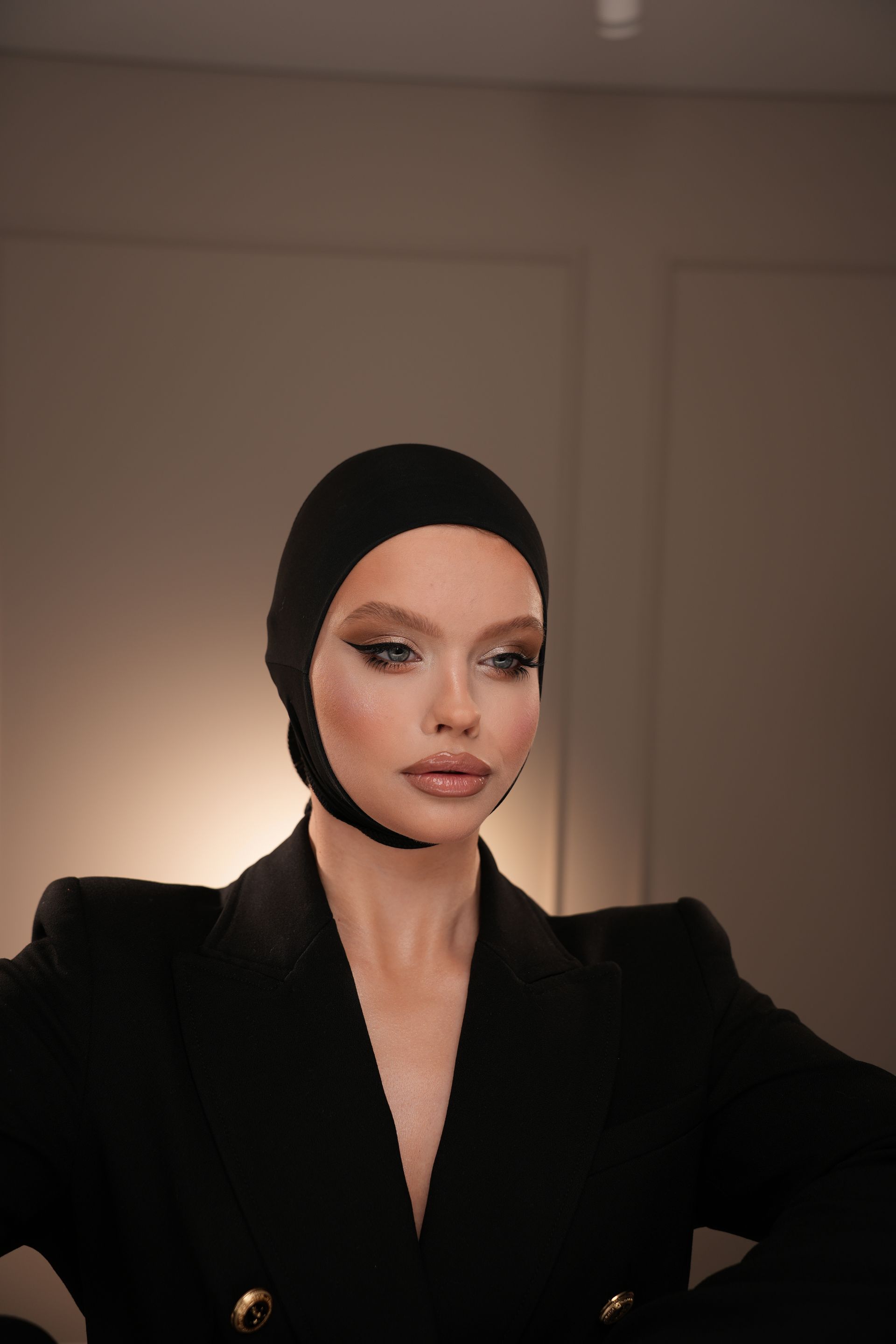Woman in black balaclava and blazer, dramatic makeup, studio setting.