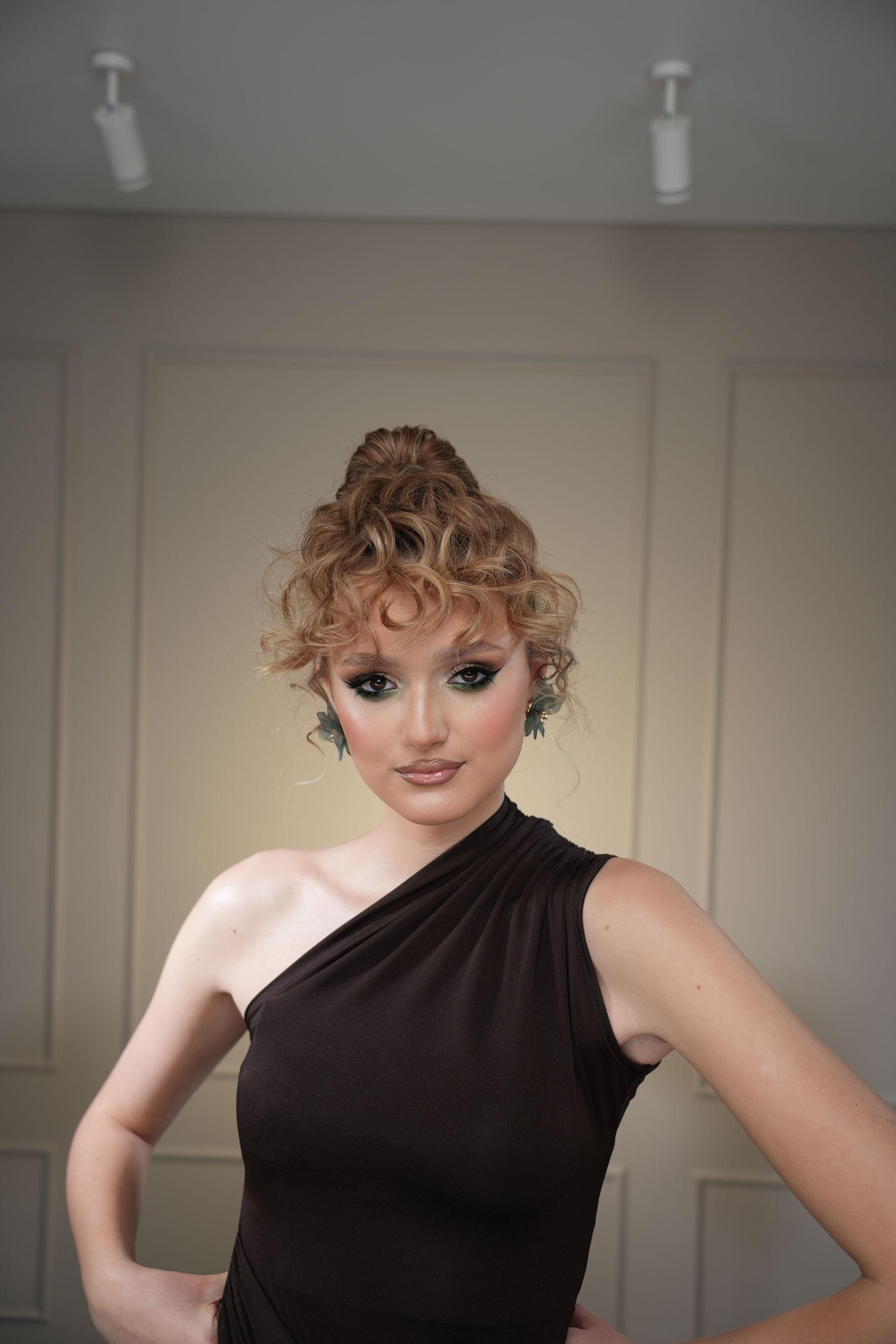 Woman with styled curly hair in a brown one-shoulder top, dark eye makeup, hands on hips. Beige wall.