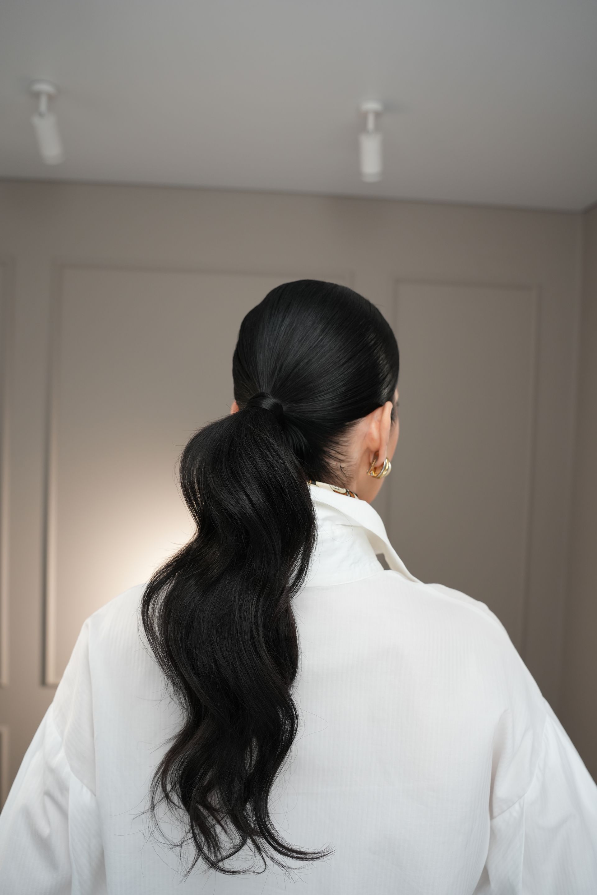 Woman in a white shirt with black hair styled in a low ponytail against a neutral background.