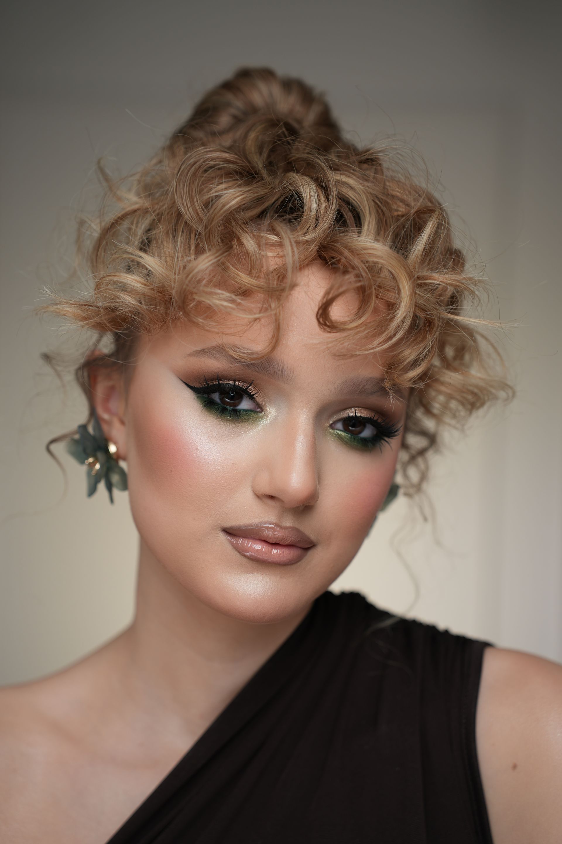 Woman with curly blonde hair styled up, wearing dramatic green eyeshadow, a brown one-shoulder top, and floral earrings.