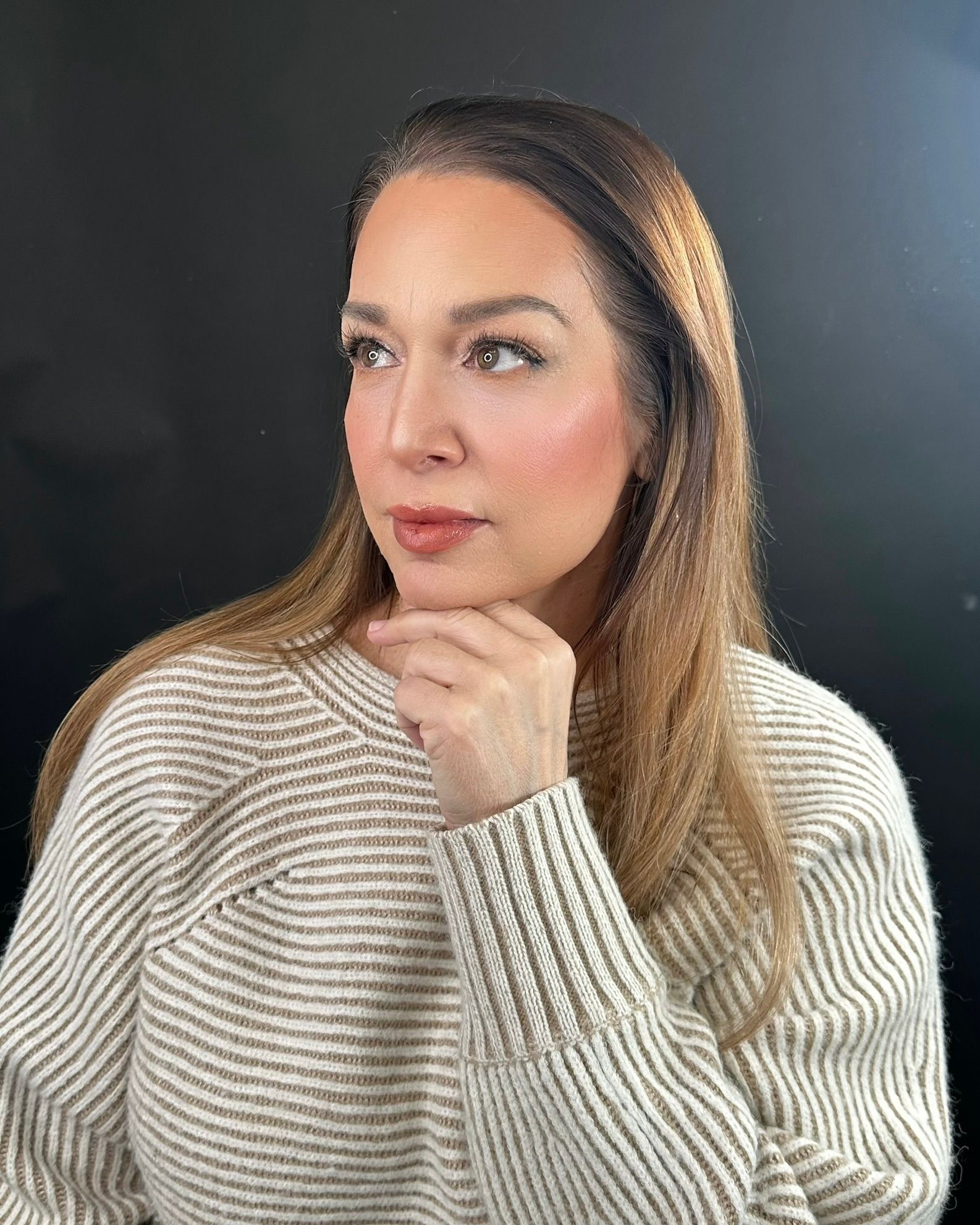 Woman in a striped sweater gazes to the left, hand on her chin, against a black backdrop.