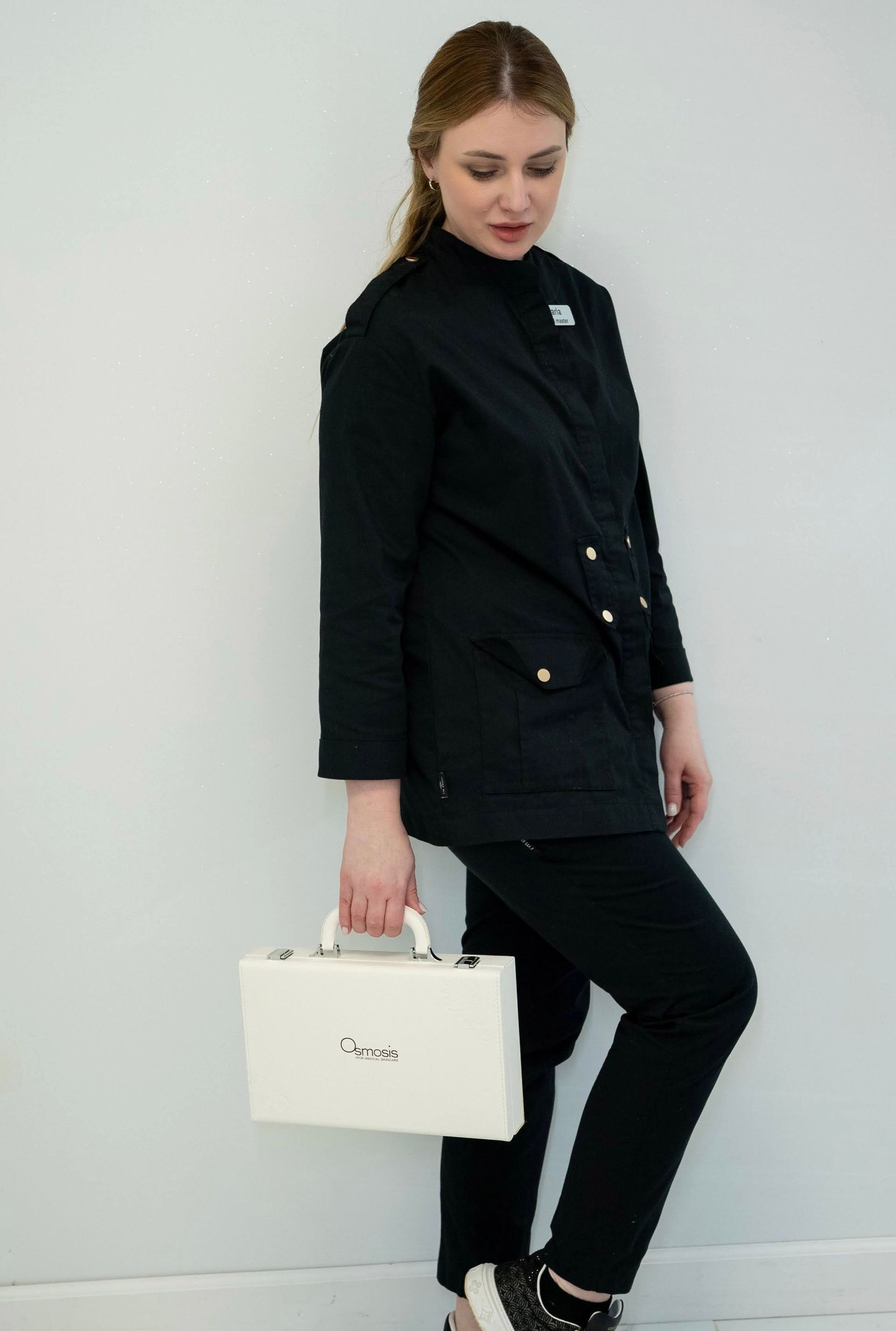 Woman in black uniform, holding a white case, leaning against a white wall.