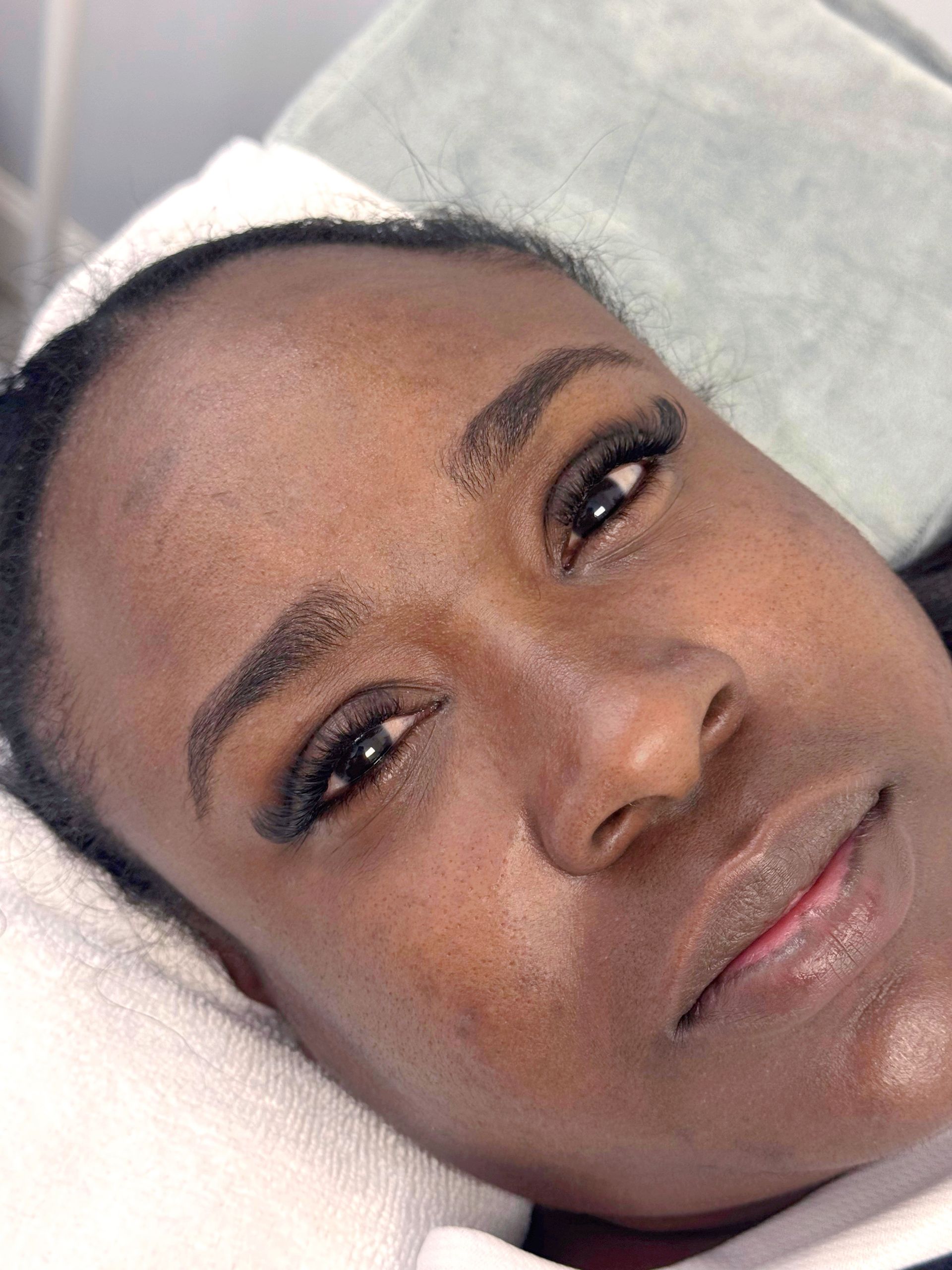 Woman with semi-permanent eyebrow makeup, resting face down on a white surface, with delicate eyelash extensions.