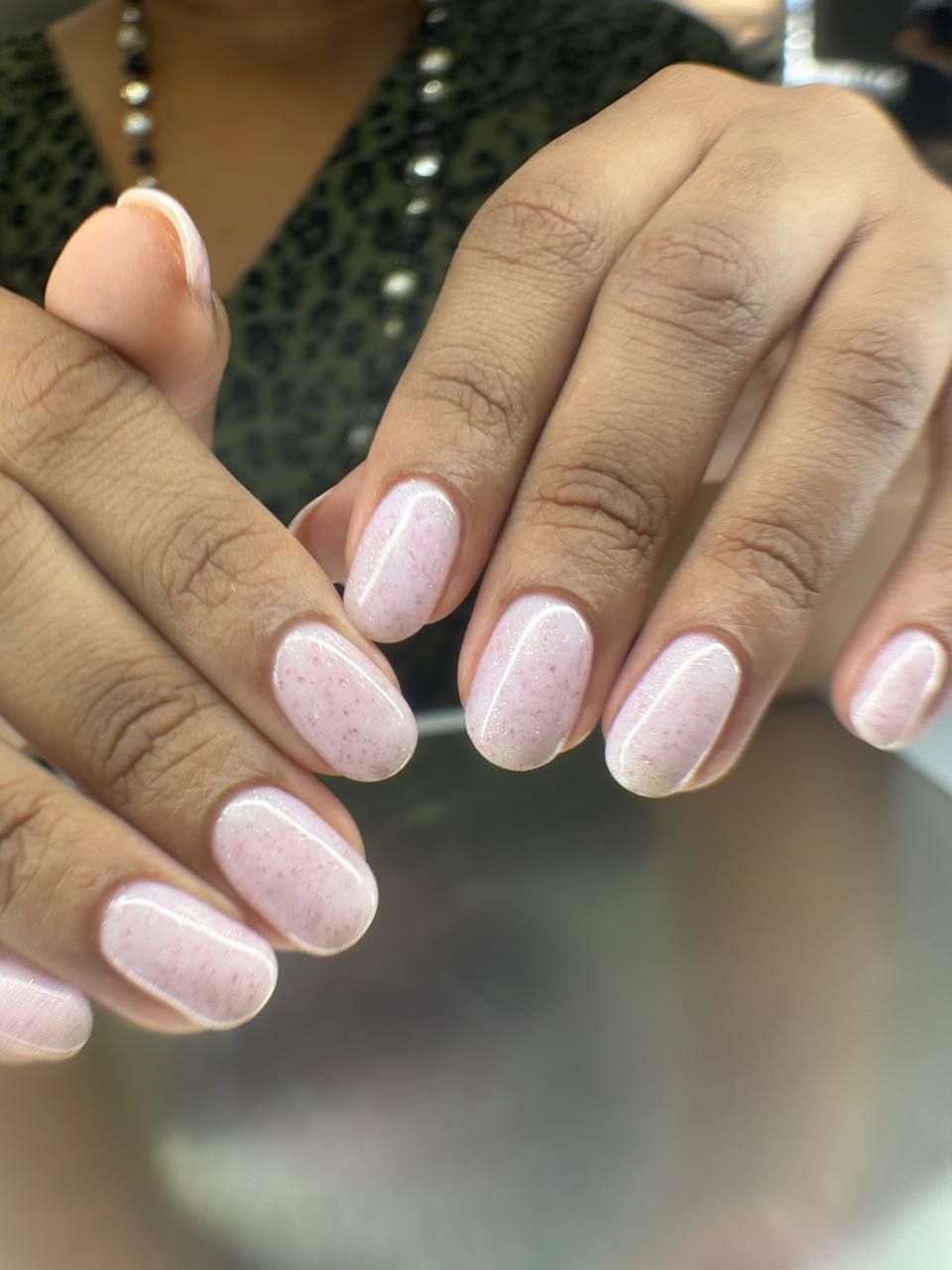 Hands with oval-shaped nails painted light pink with sparkles, in front of a green top.