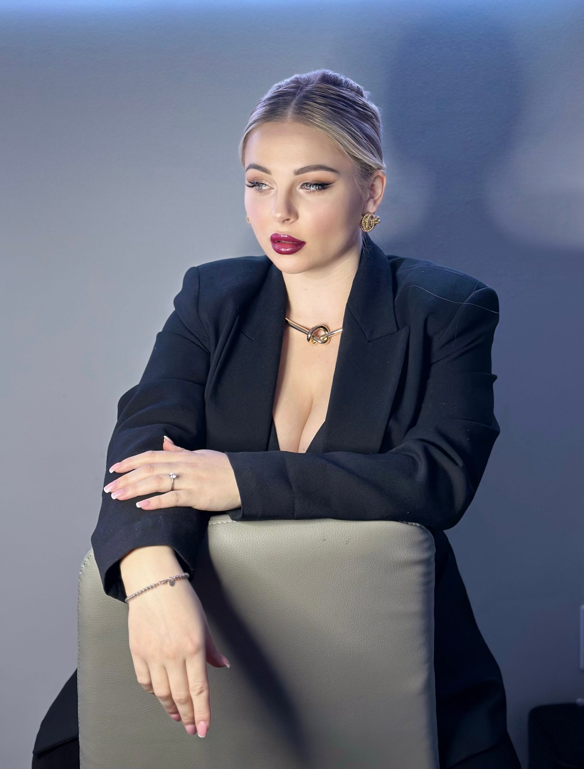 Woman in black blazer, leaning on a chair, looking left with a serious expression.