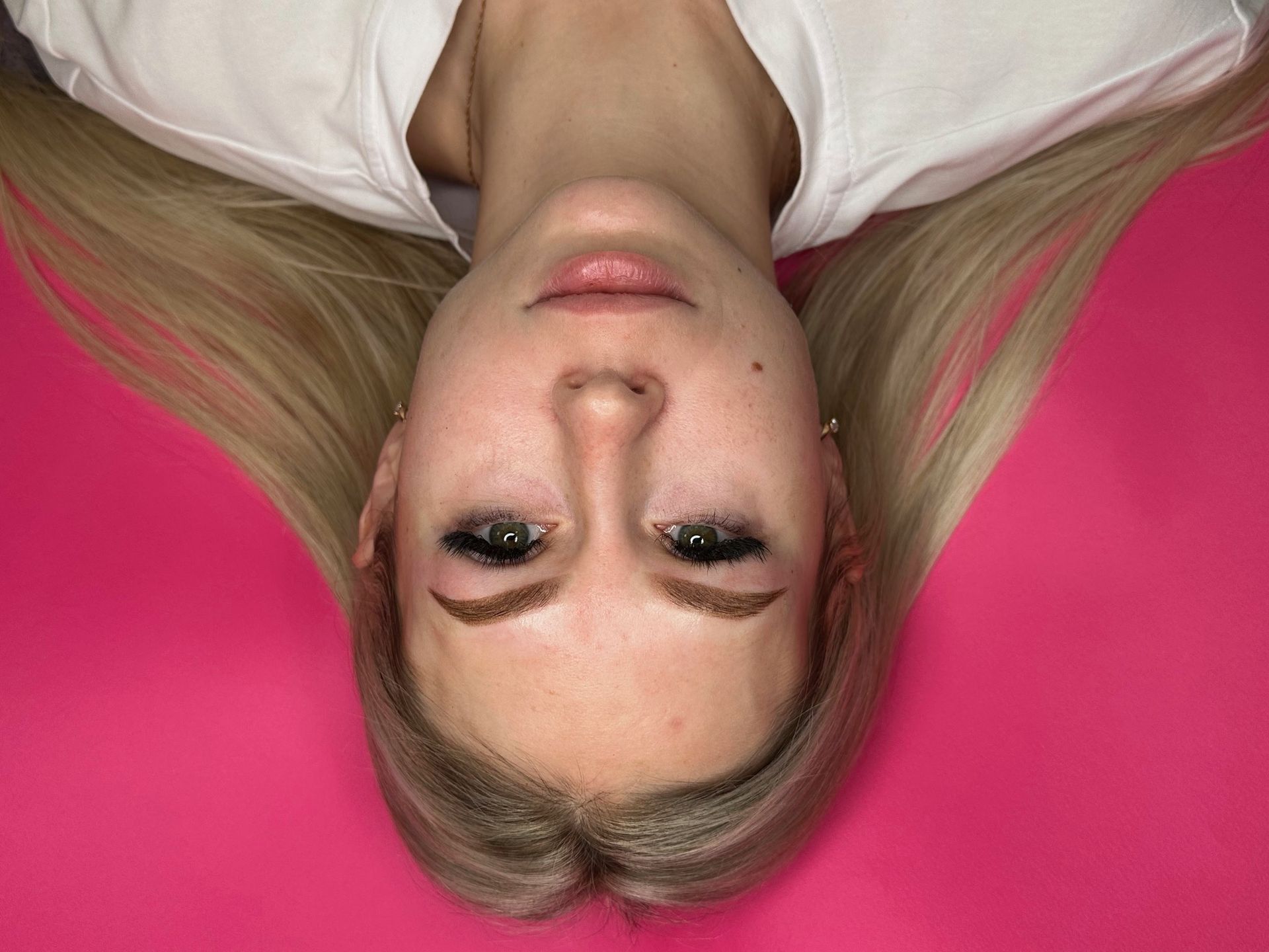 Woman with blonde hair and eye makeup, lying on a pink surface with a white shirt.