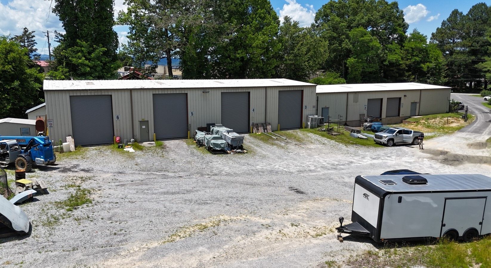 Metal buildings with garage doors, vehicles, and a trailer on a gravel lot; trees in the background.