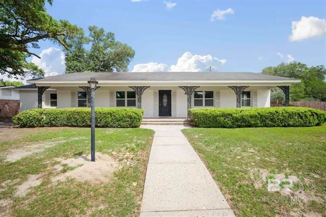 Single-story beige house with a front walkway, trimmed hedges, and a lawn under a bright sky