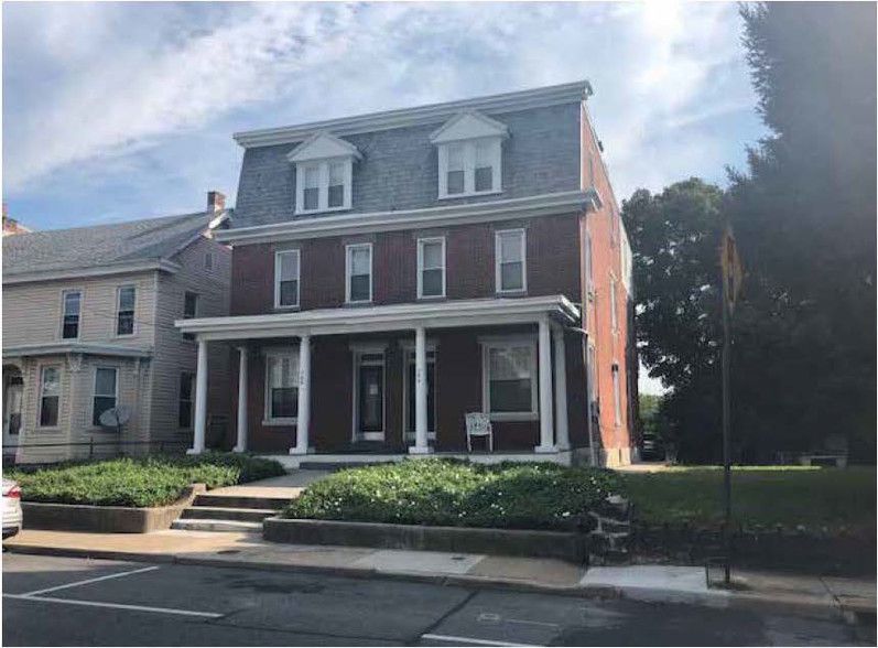 Three-story brick house with a grey mansard roof, white porch, and front yard with bushes.