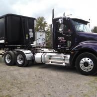 A dark purple semi-truck with a black roll-off dumpster trailer parked on a gravel lot under a cloudy sky.
