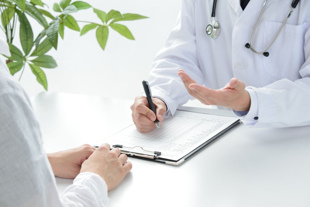Doctor writing on a clipboard while speaking with a patient at a bright office desk — Tewantin Medical Centre In Tewantin, QLD