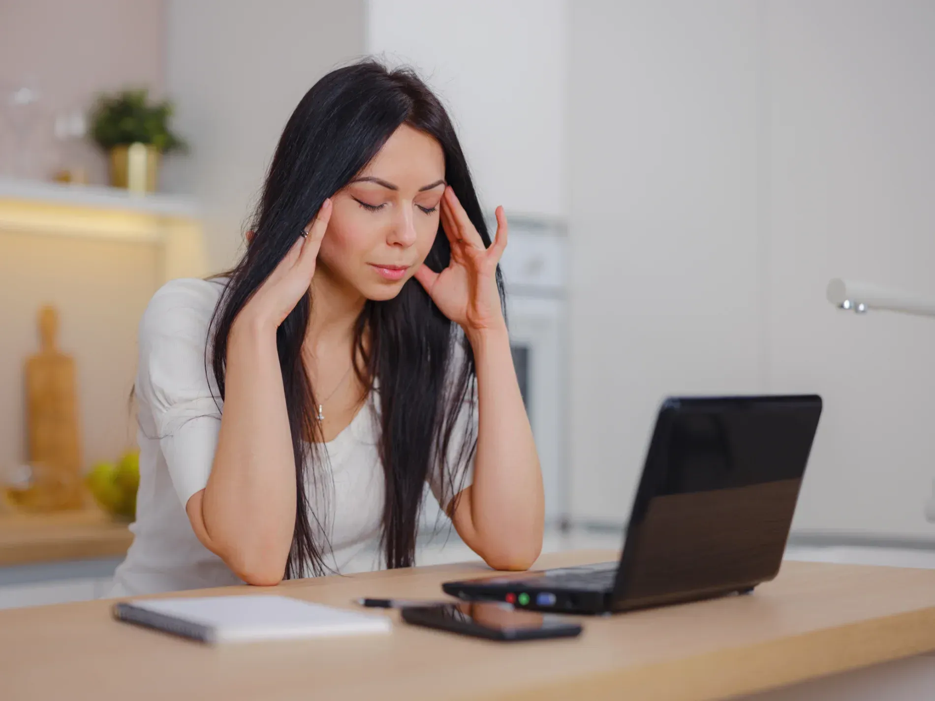 Woman at laptop, touching temples, appearing tired, in a kitchen setting  — Tewantin Medical Centre In Tewantin, QLD