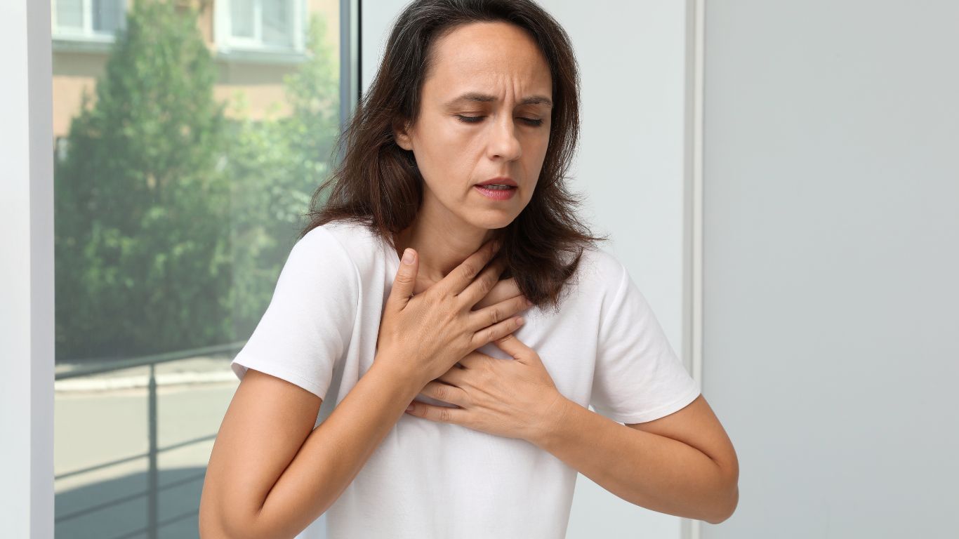 Woman in White Shirt, Hands on Chest, Struggling to Breathe, by a Window — Tewantin Medical Centre In Tewantin, QLD