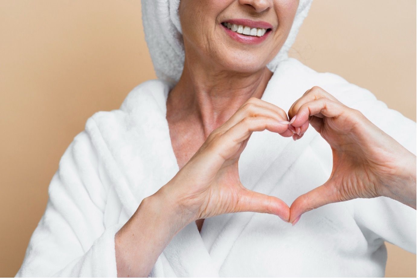 Woman in white robe and towel making a heart shape with her hands, smiling.