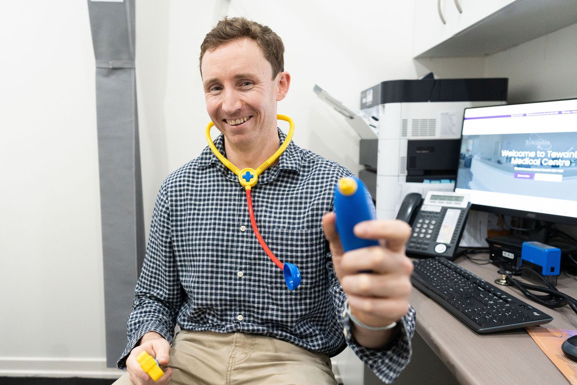 Doctor smiling, holding a toy stethoscope and medical instrument in an office setting.