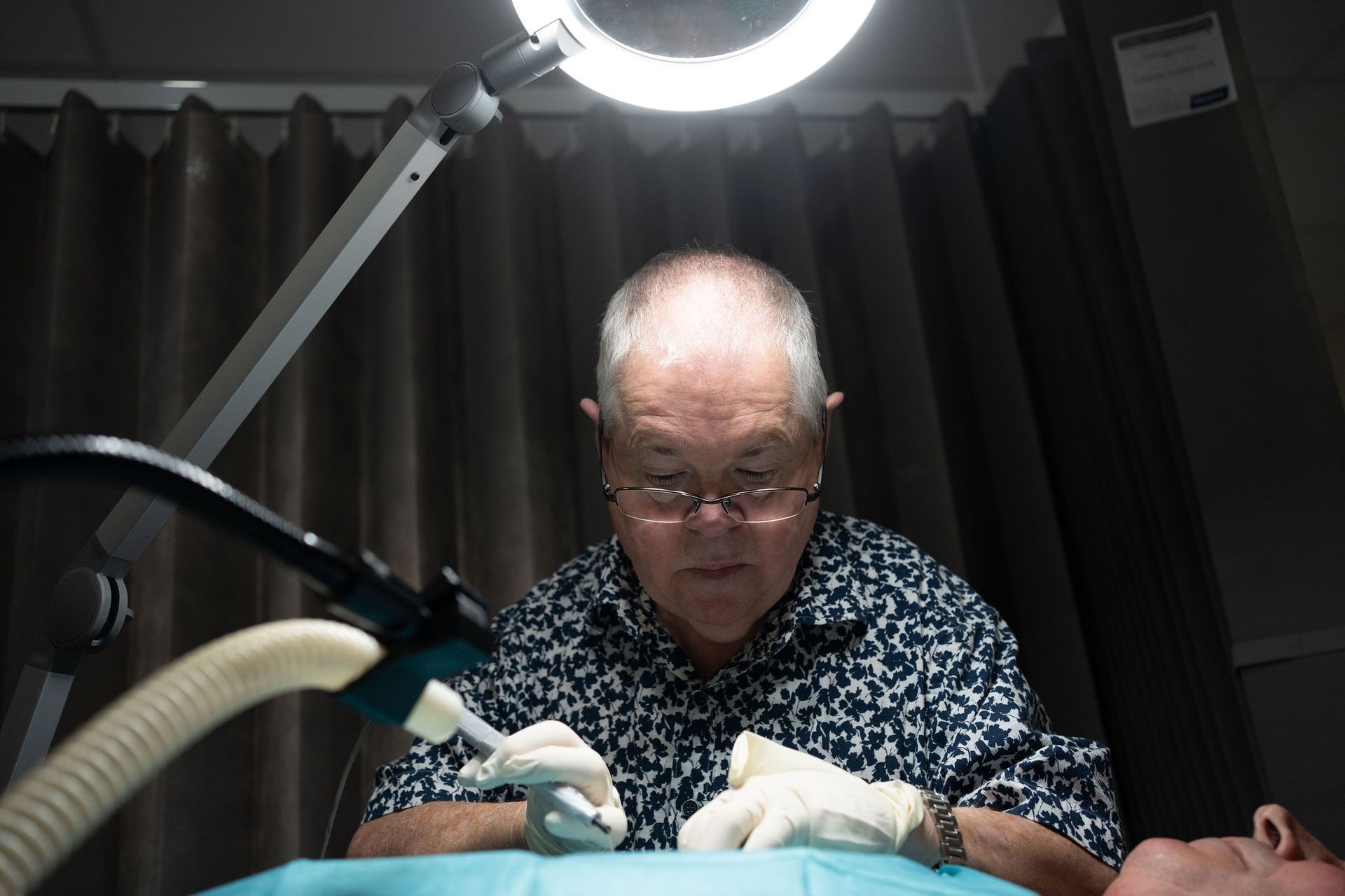 Doctor performing a procedure under a magnifying lamp. He wears gloves and glasses.