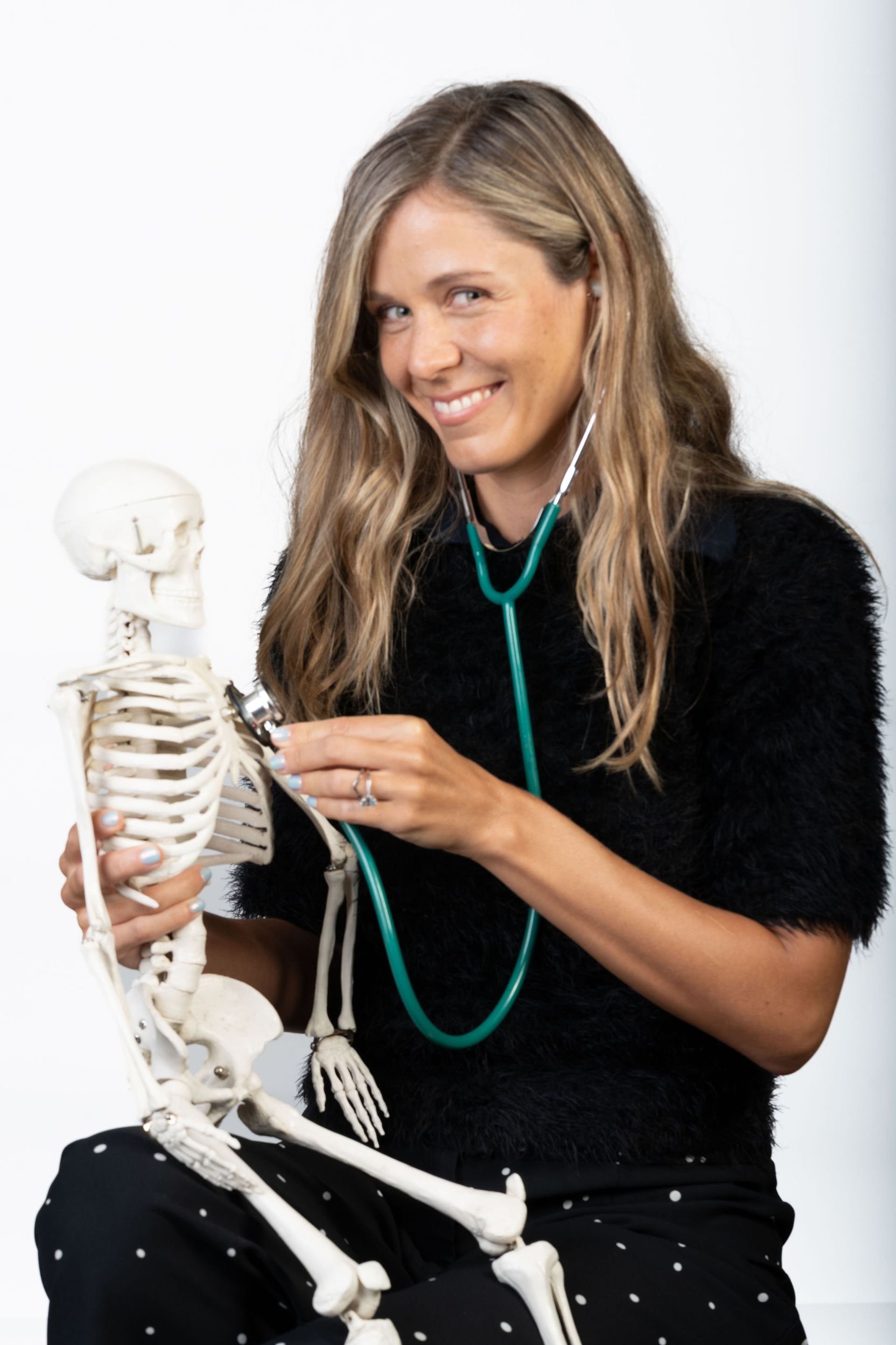 Woman with stethoscope examining a skeleton, smiling; white background.