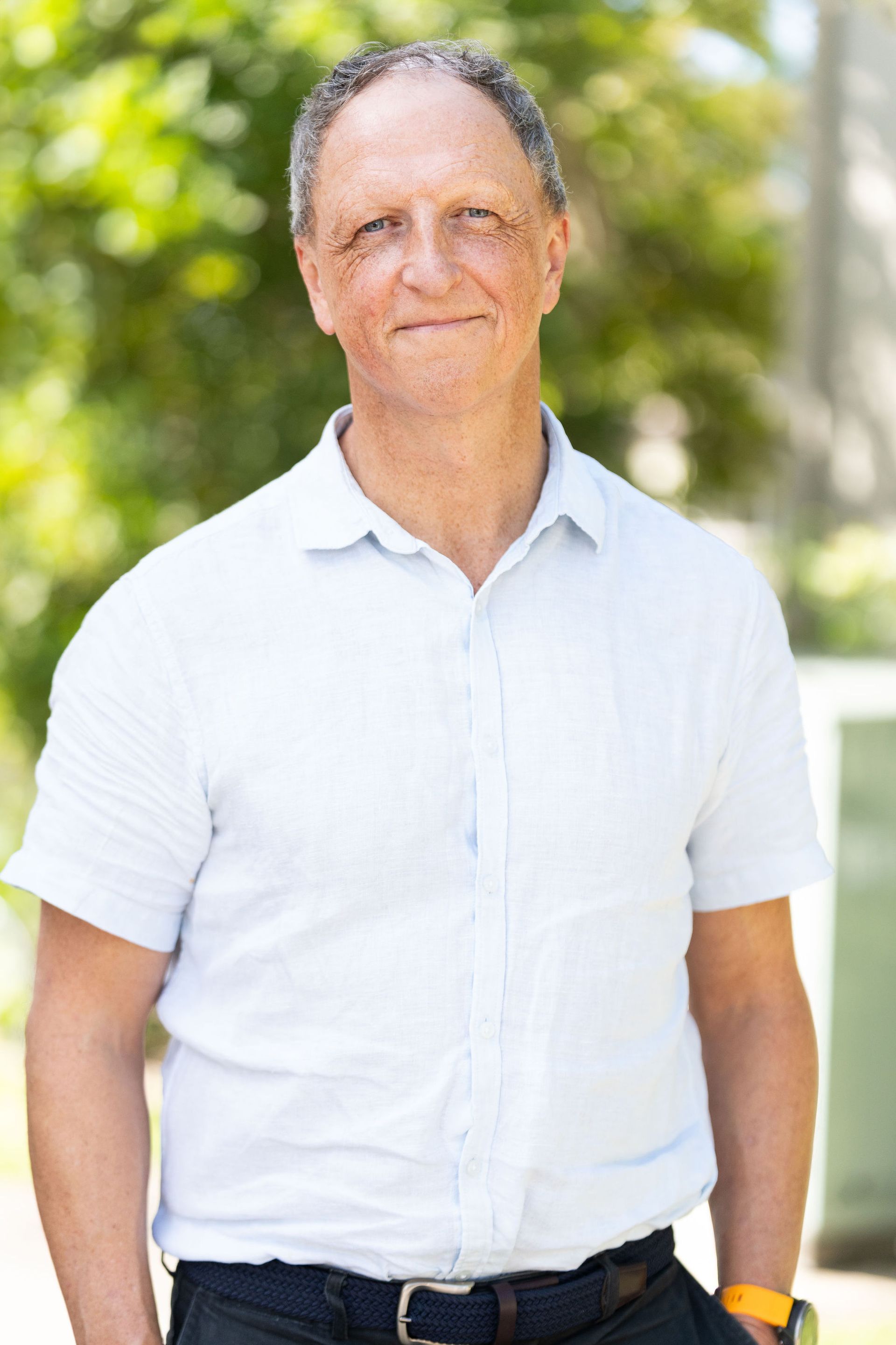 Man in a white shirt and dark pants smiles outdoors.