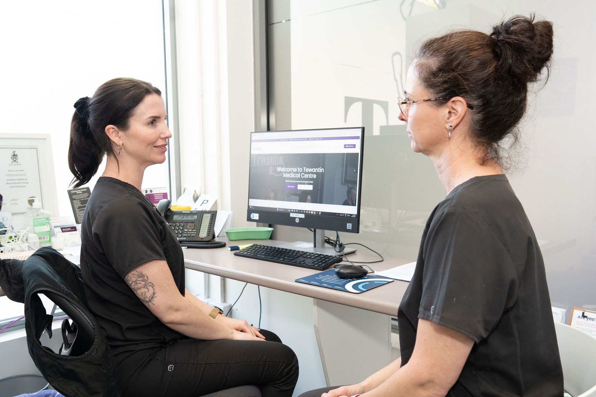 Two women in black scrubs consult at a computer in a brightly lit office.
