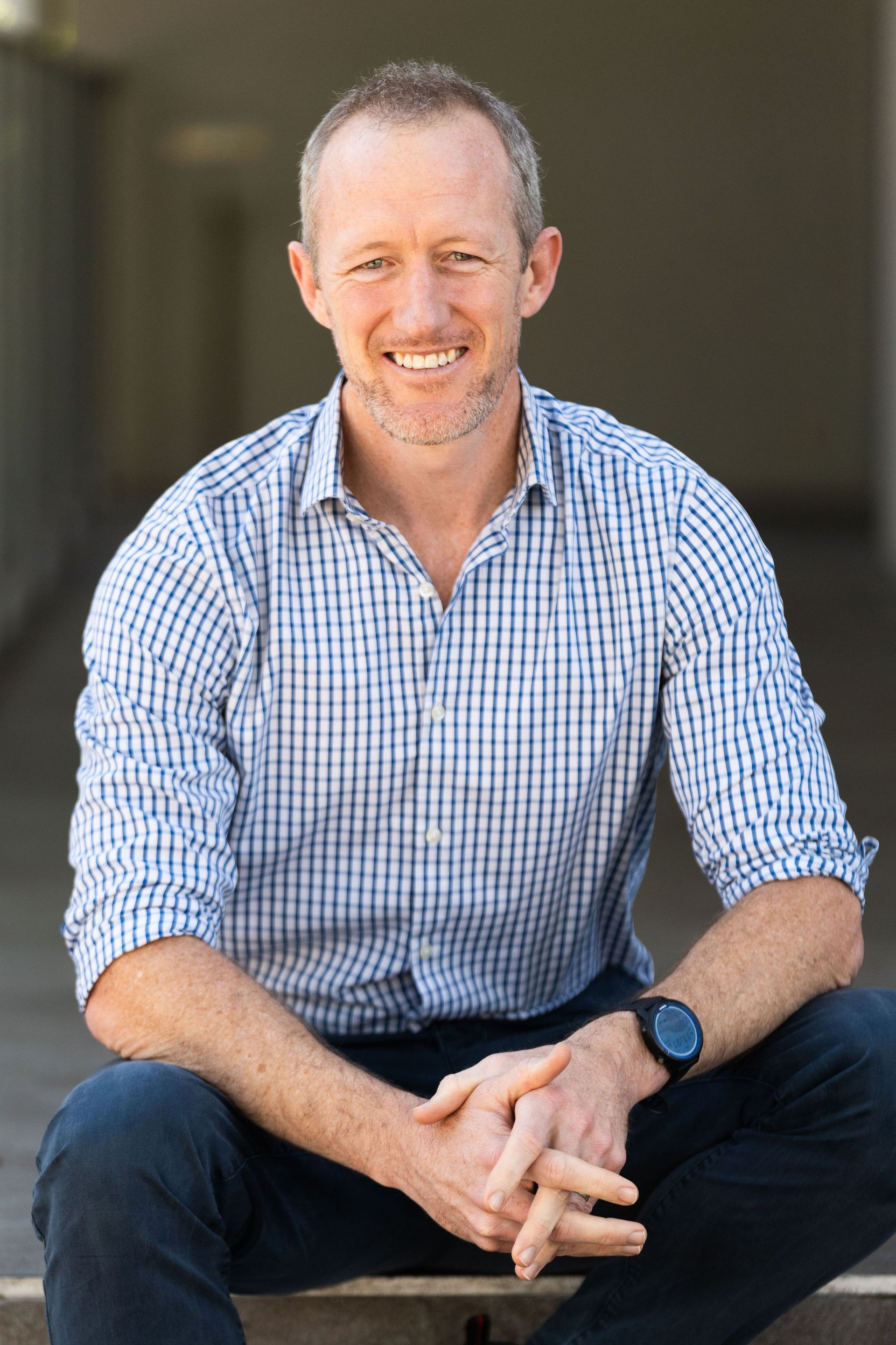 Man smiling, sitting outdoors, wearing blue plaid shirt and dark pants, hands clasped.