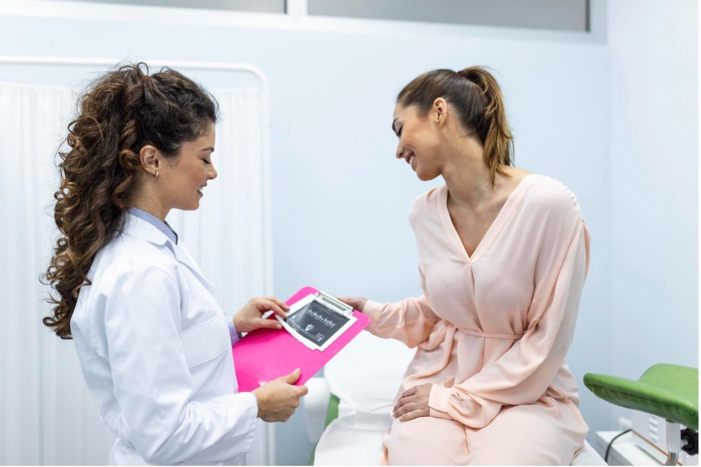 Doctor showing ultrasound to a woman in a medical office; woman smiles, sitting on a medical bed.