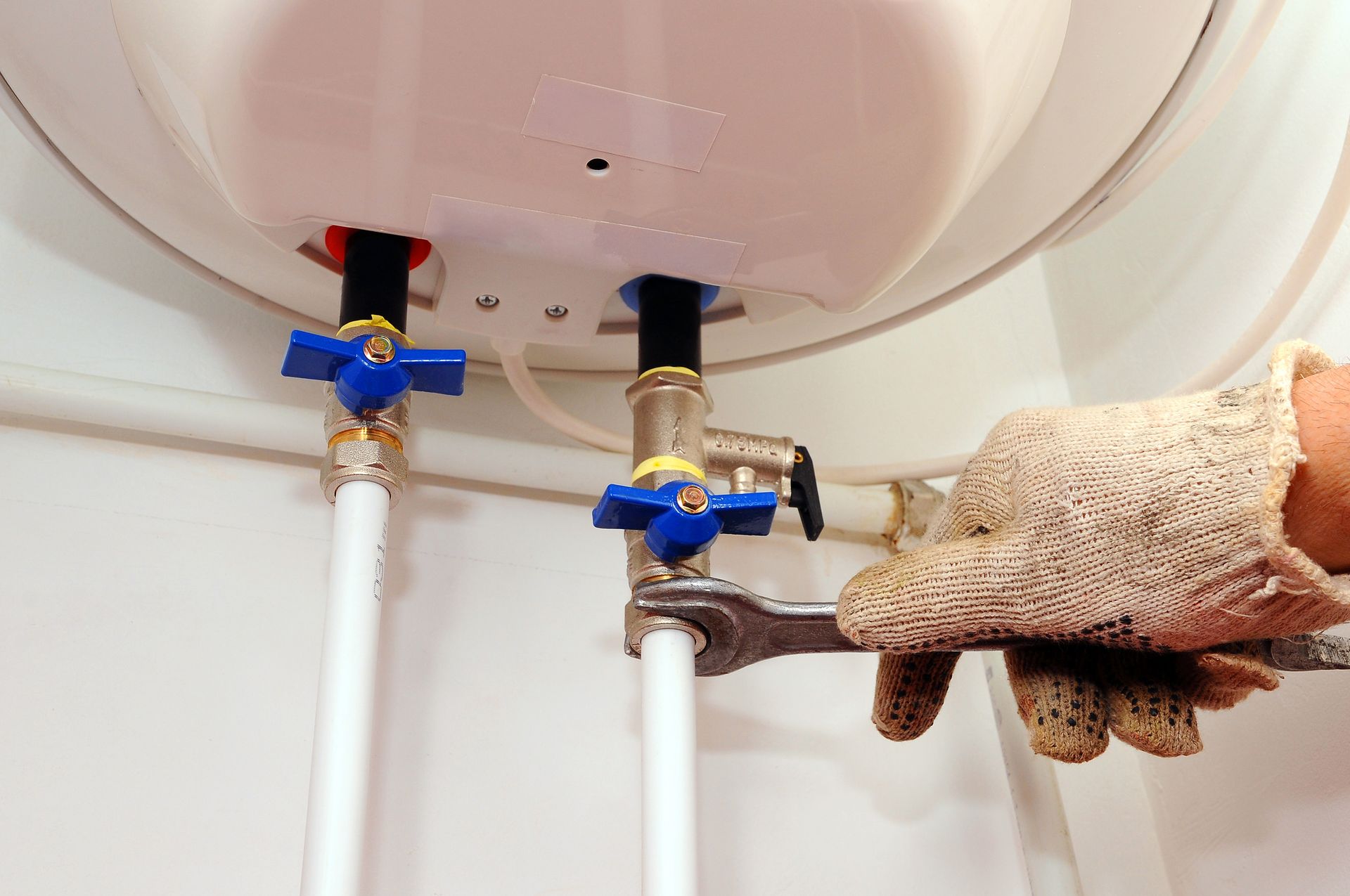 Person in work gloves using a wrench to work on a hot water tank with blue handled valves and white pipes. Person in work gloves using a wrench to work on a hot water tank with blue handled valves and white pipes.