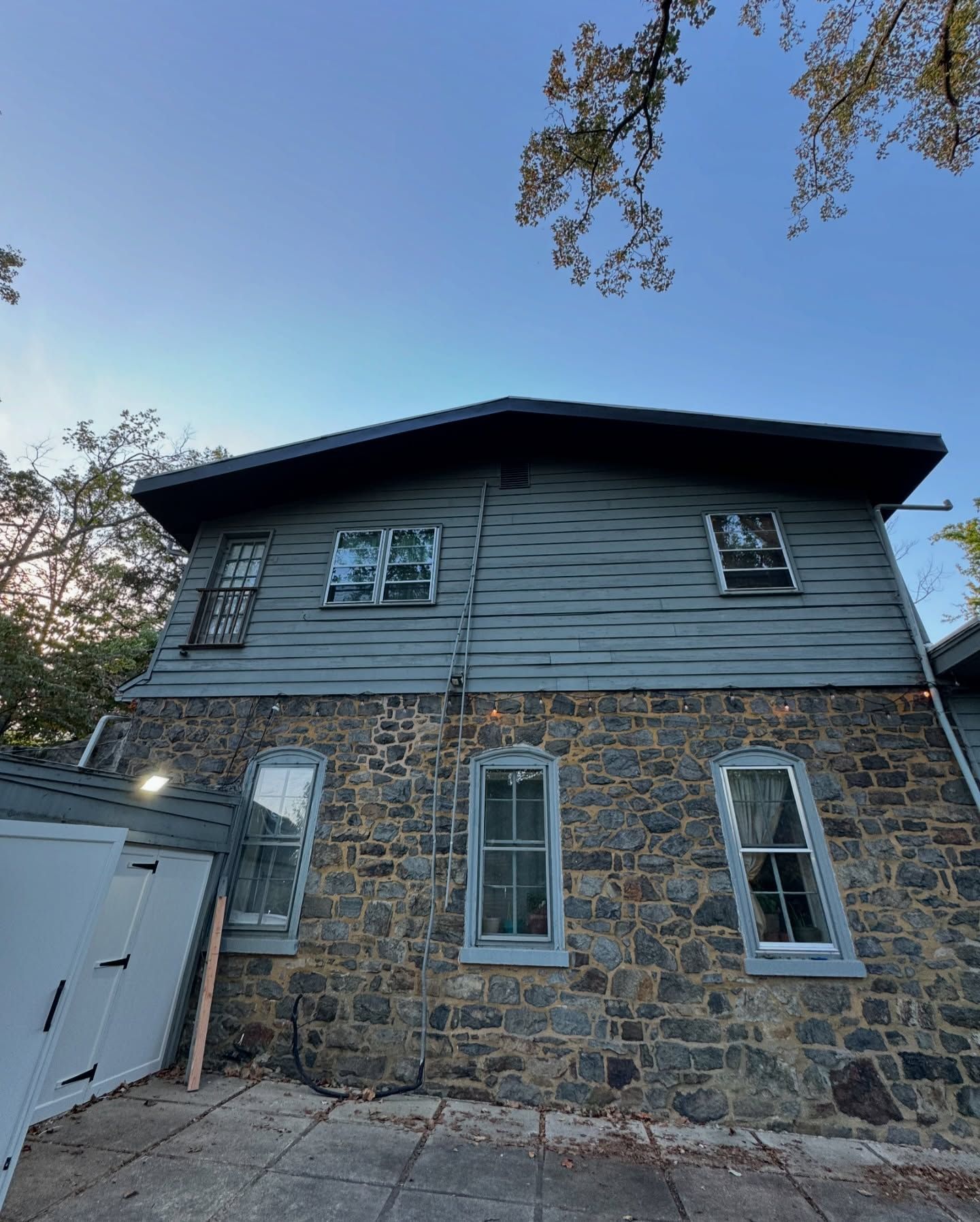 Two-story house with stone bottom and gray siding top. Windows are visible. Overcast sky.