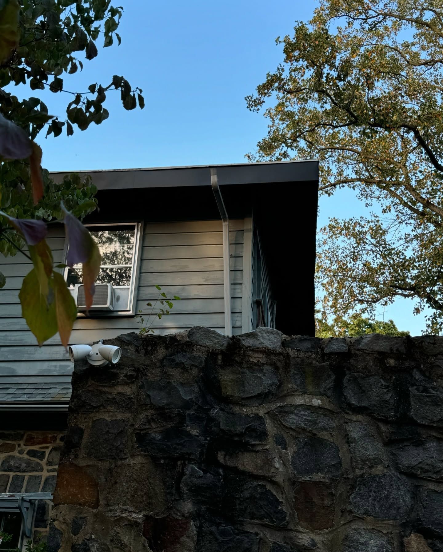 Gray house with stone wall, trees, and blue sky.