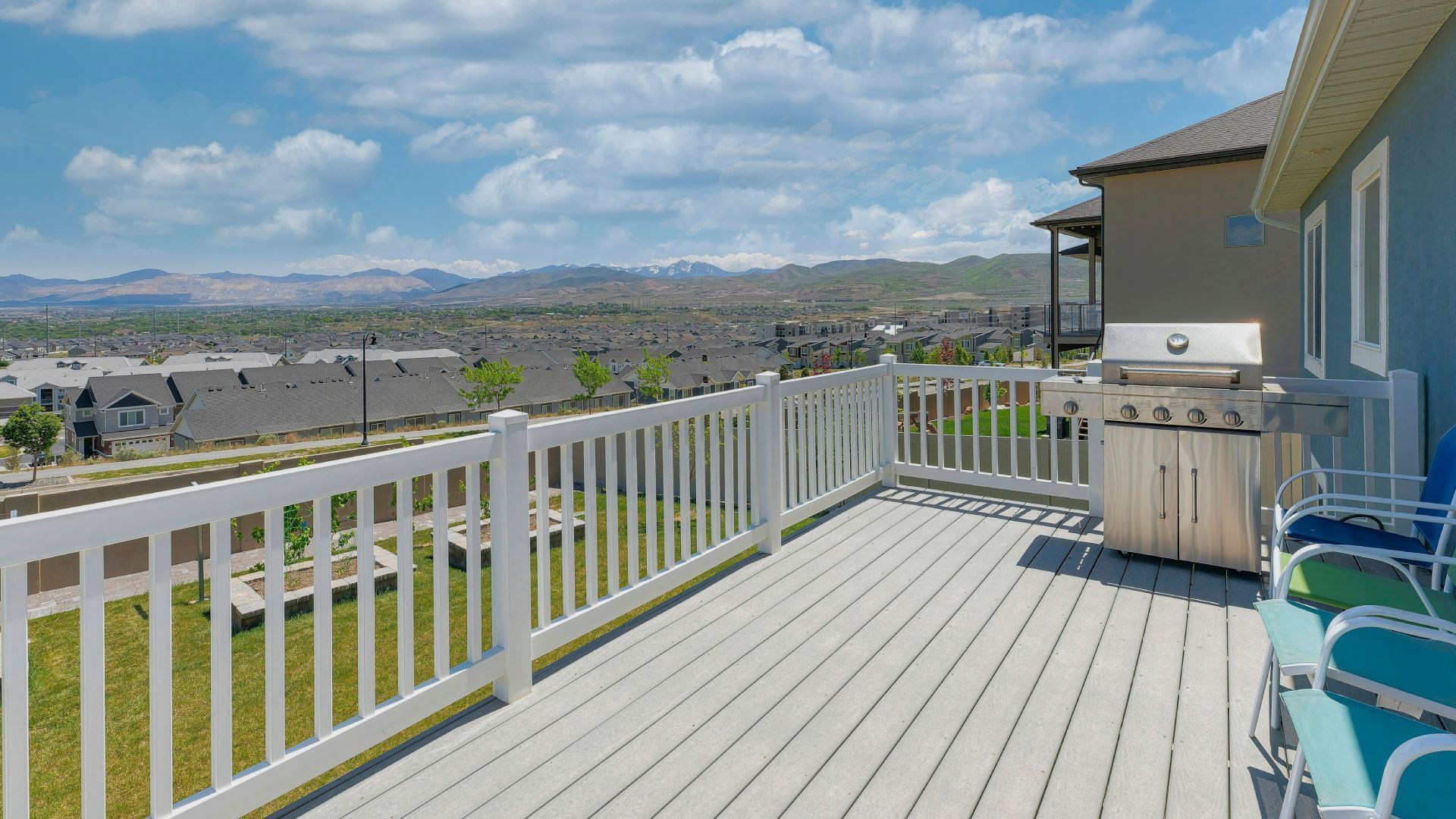Deck with white railing, grill, and blue chairs, overlooking a suburban landscape under a blue sky.
