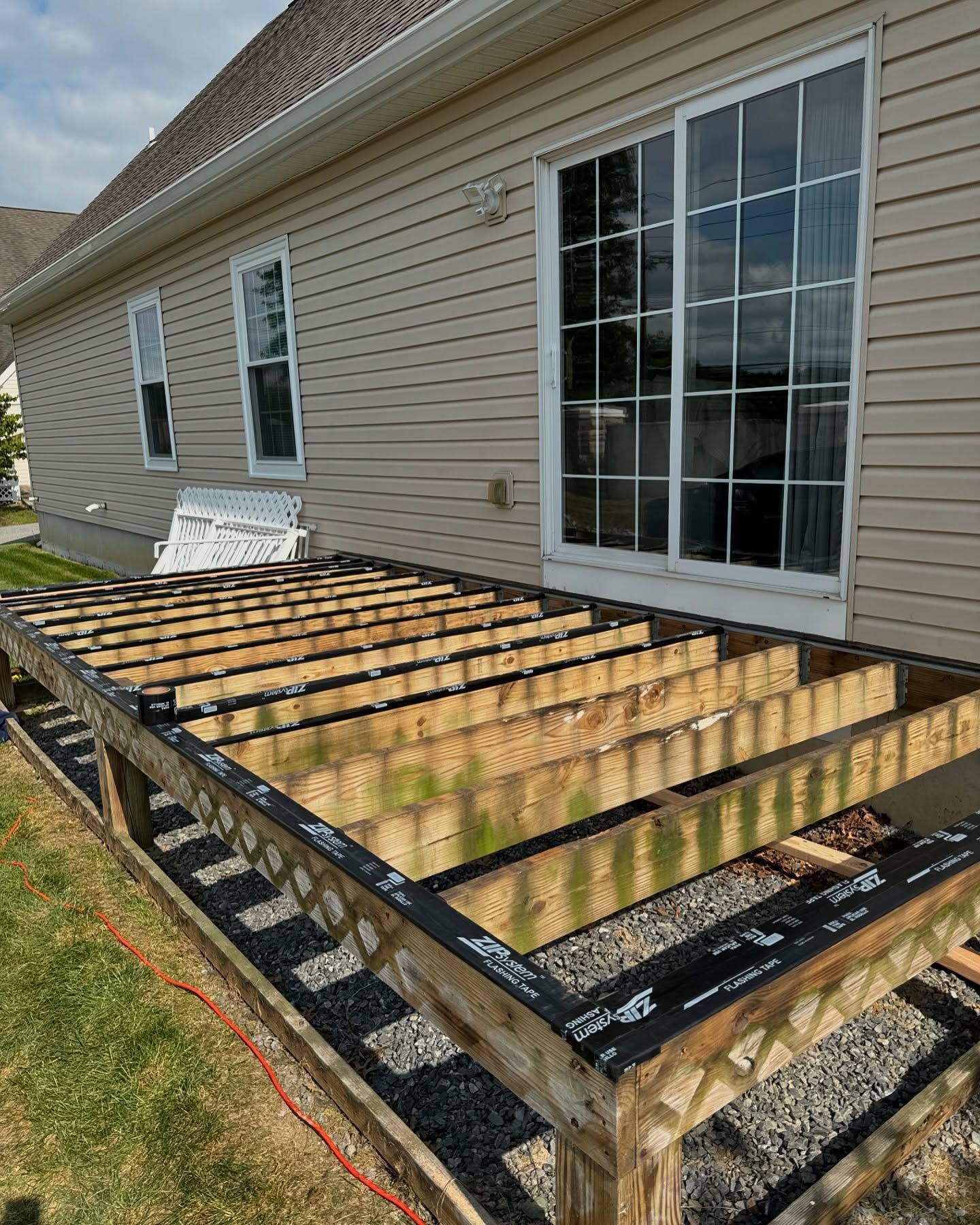 Partially constructed deck frame attached to a house with sliding glass doors. Green grass and gravel are visible.