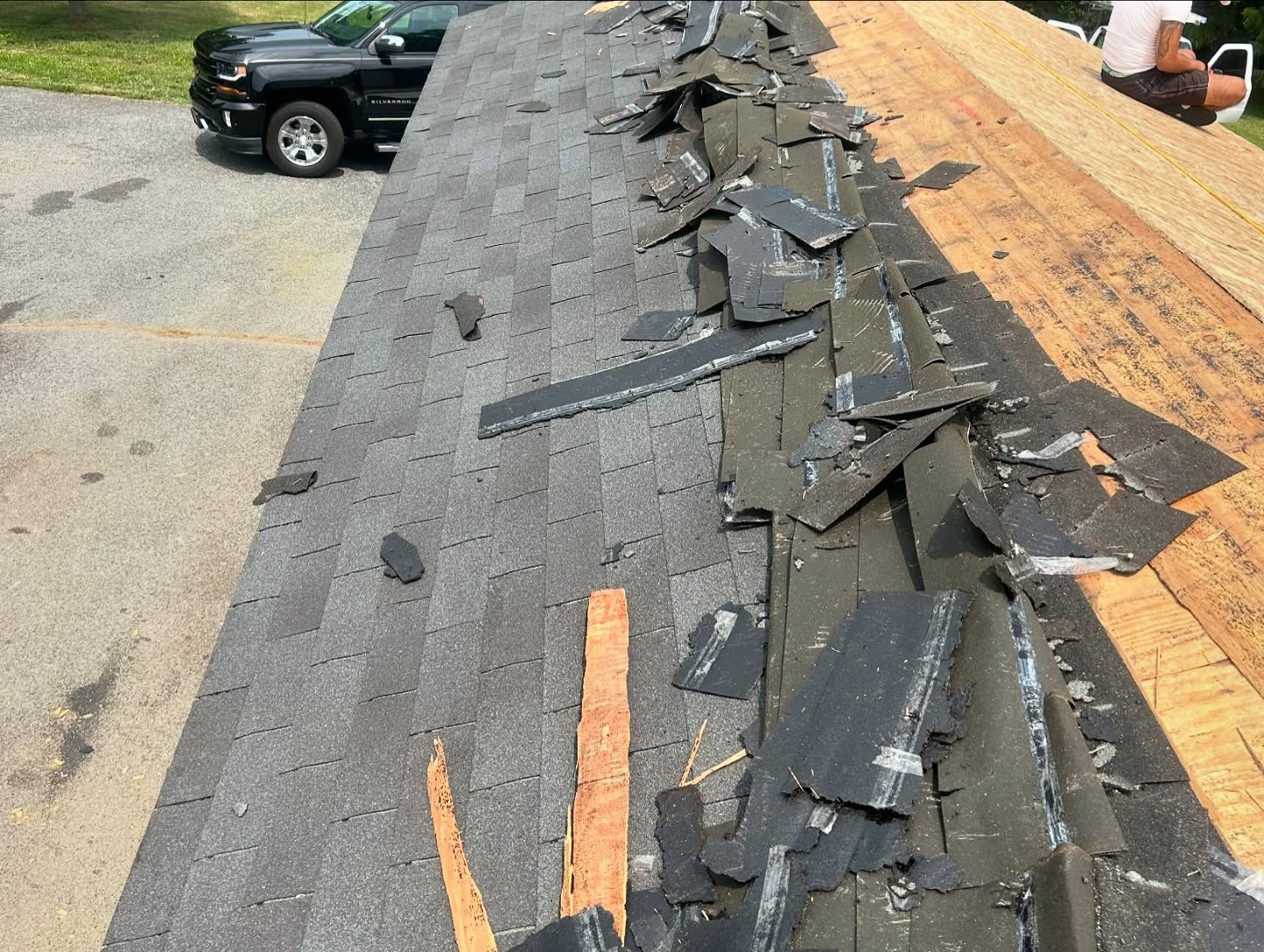 Damaged asphalt shingles on a roof during repair; black truck in background.