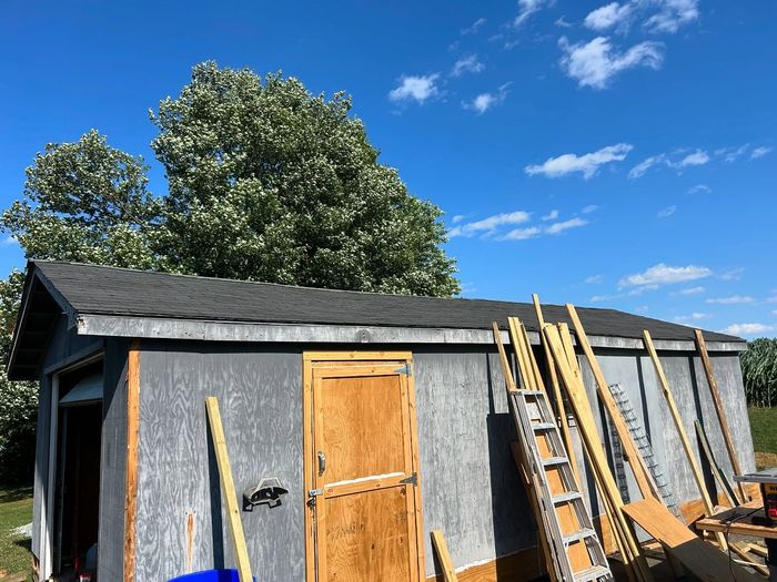 Gray shed with a dark roof and wooden door under a blue sky, with wood leaning against it.