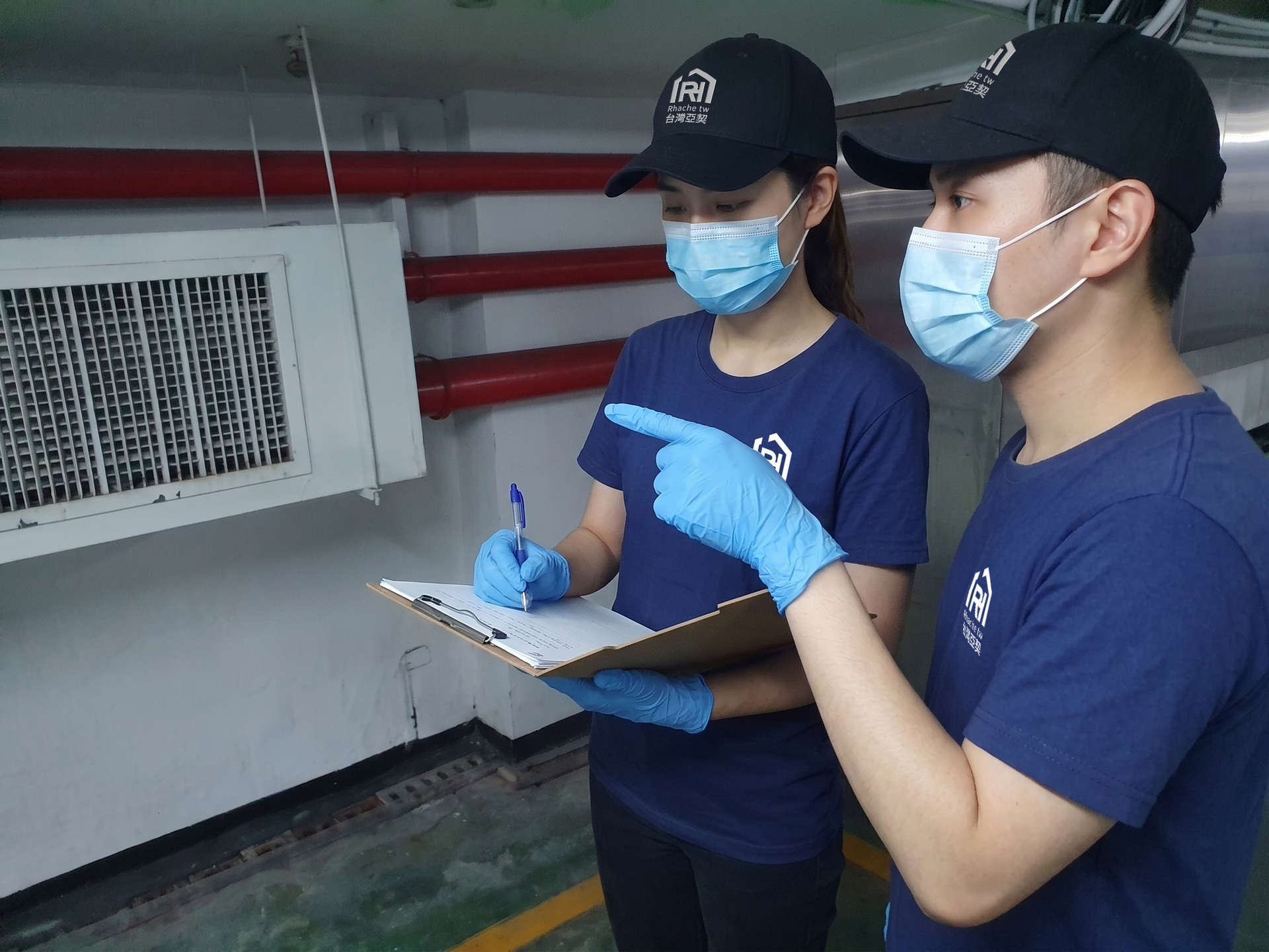 兩名身穿藍色制服、口罩的工人在工業設備旁做筆記。