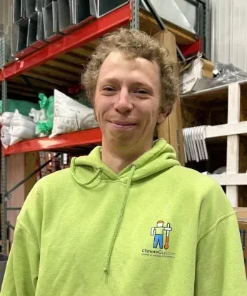 Man with curly hair in a green hoodie smiles in a warehouse setting, shelves in the background.