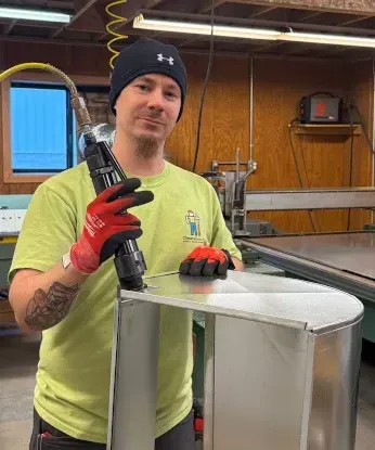 Man wearing a black beanie and red gloves working with metal in a workshop. He is using a tool on a curved metal structure.