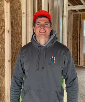 Man in red hat and gray hoodie stands inside a building frame, smiling. He has a logo on the hoodie.