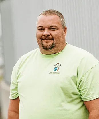 A middle-aged man with a beard wearing a green t-shirt smiles at the camera outdoors. The shirt has a small logo on the chest.