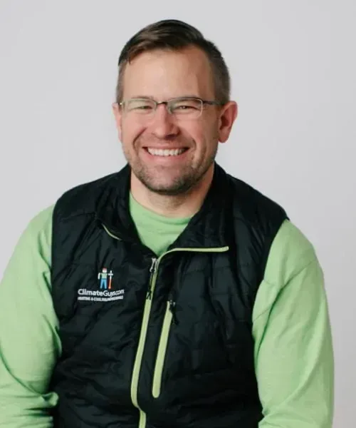 Smiling man in glasses, green shirt, and black vest with logo, against a white background.