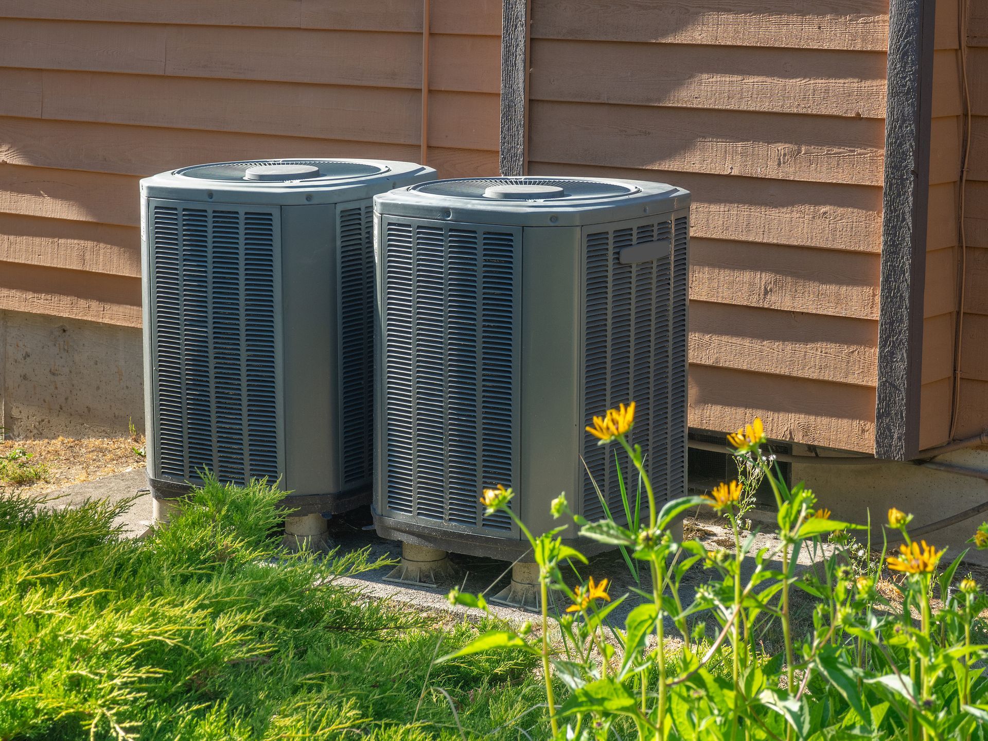 Two gray air conditioning units are positioned outside a wooden building, surrounded by greenery and yellow wildflowers.